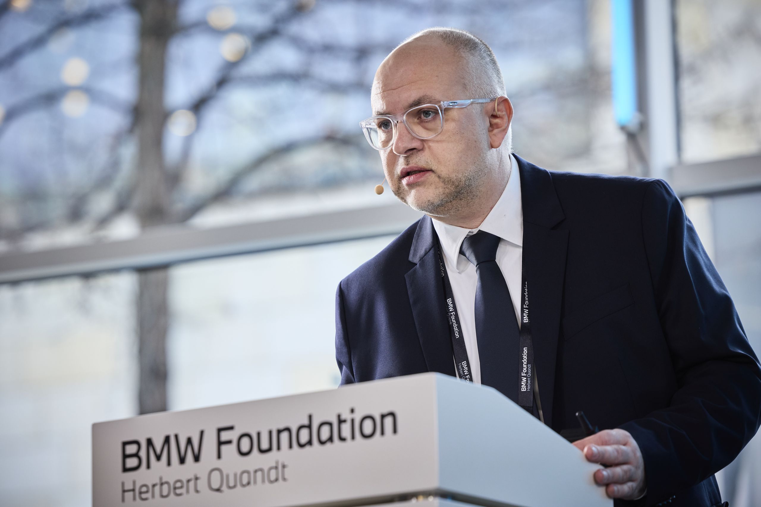 A man in a suit speaks at a podium with "BMW Foundation Herbert Quandt" written on it, in a modern indoor setting with natural light.