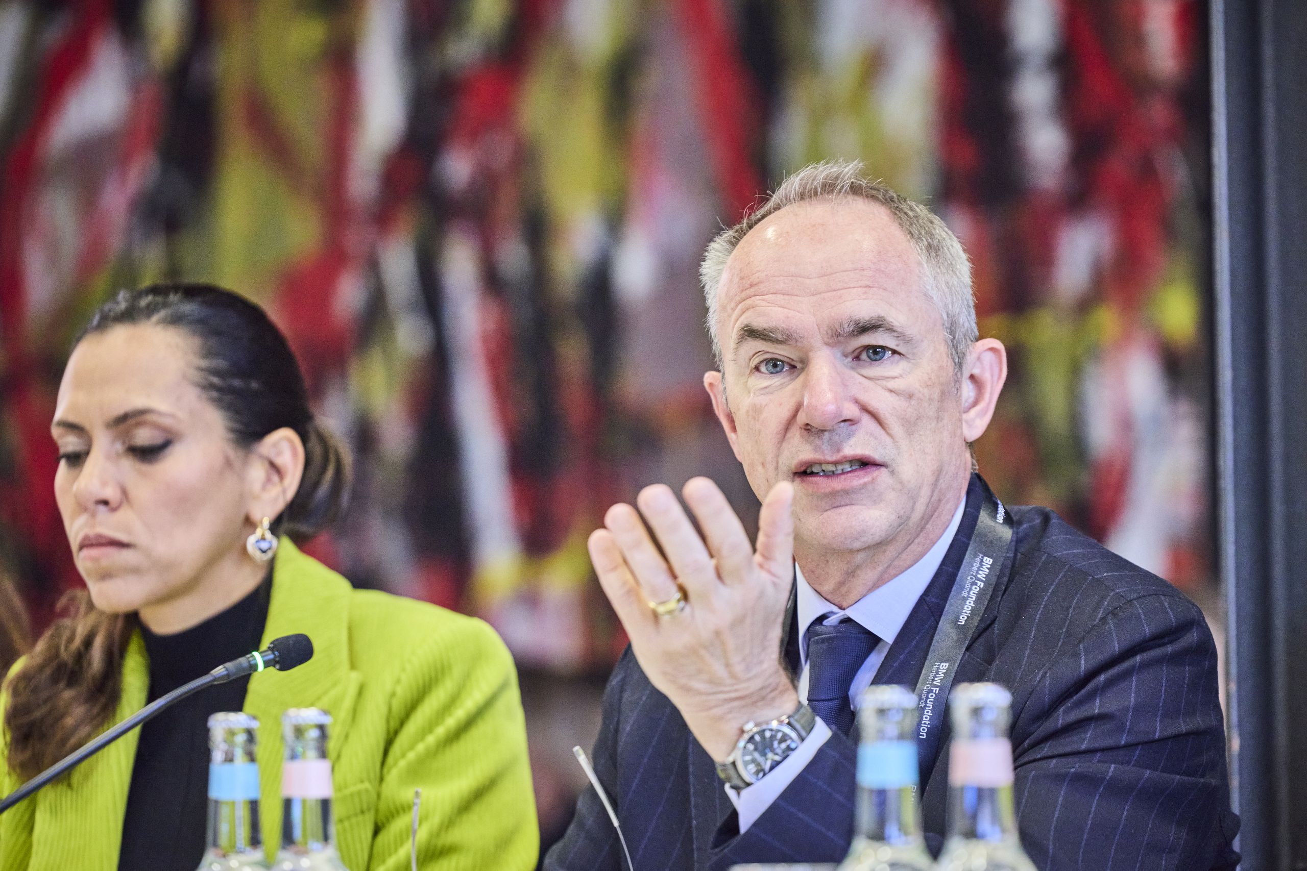 Heba Aguib, Member of the Board, BMW Foundation Herbert Quandt and Jean-Christophe Laloux, Director General – Head of EU Lending and Advisory Operations at the European Investment Bank (EIB). A man in a suit gestures while speaking at a conference, seated next to a woman in a green jacket. Bottles are on the table in front of them.