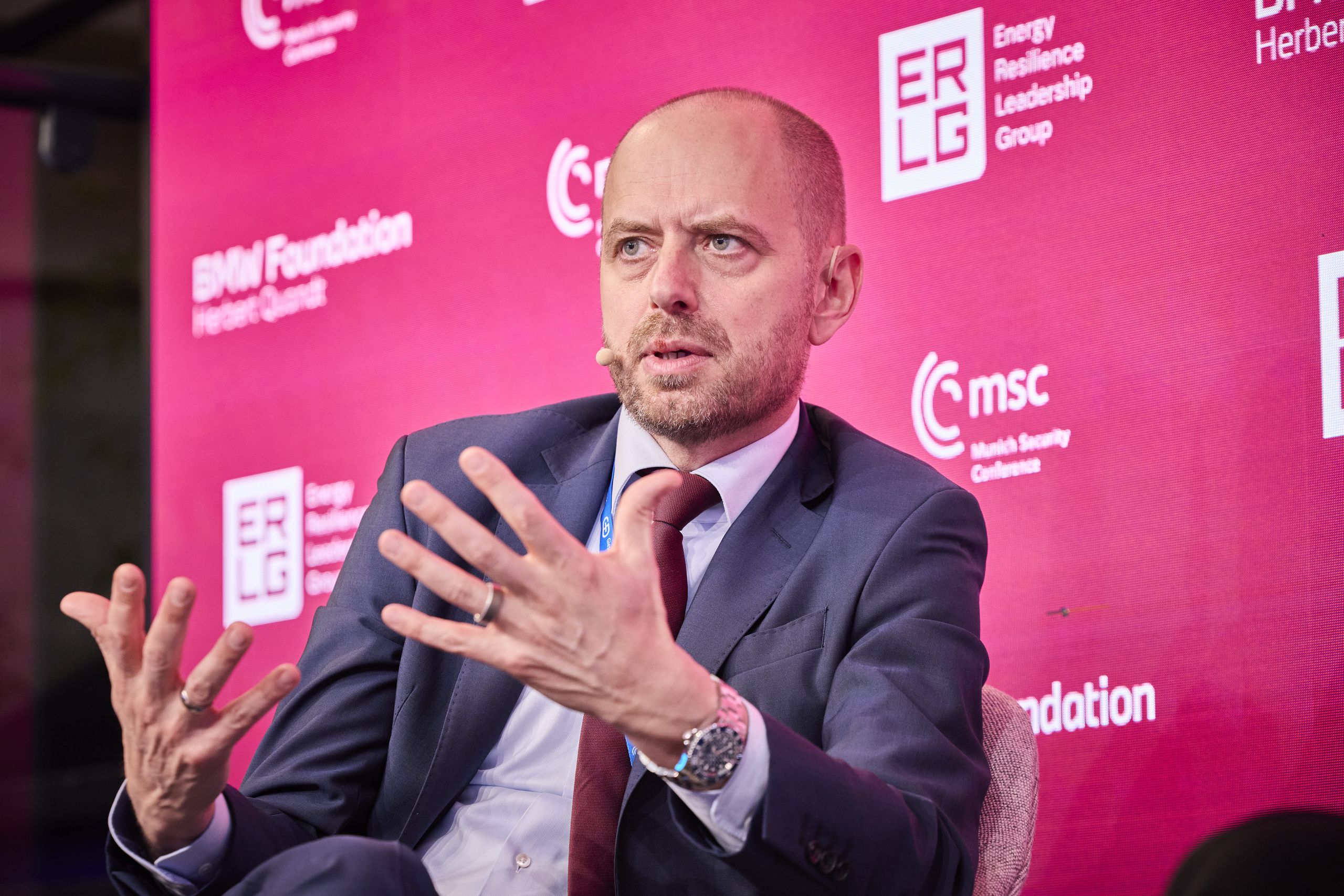 Man in a suit speaking animatedly, seated against a pink backdrop with text and logos, wearing a microphone headset.