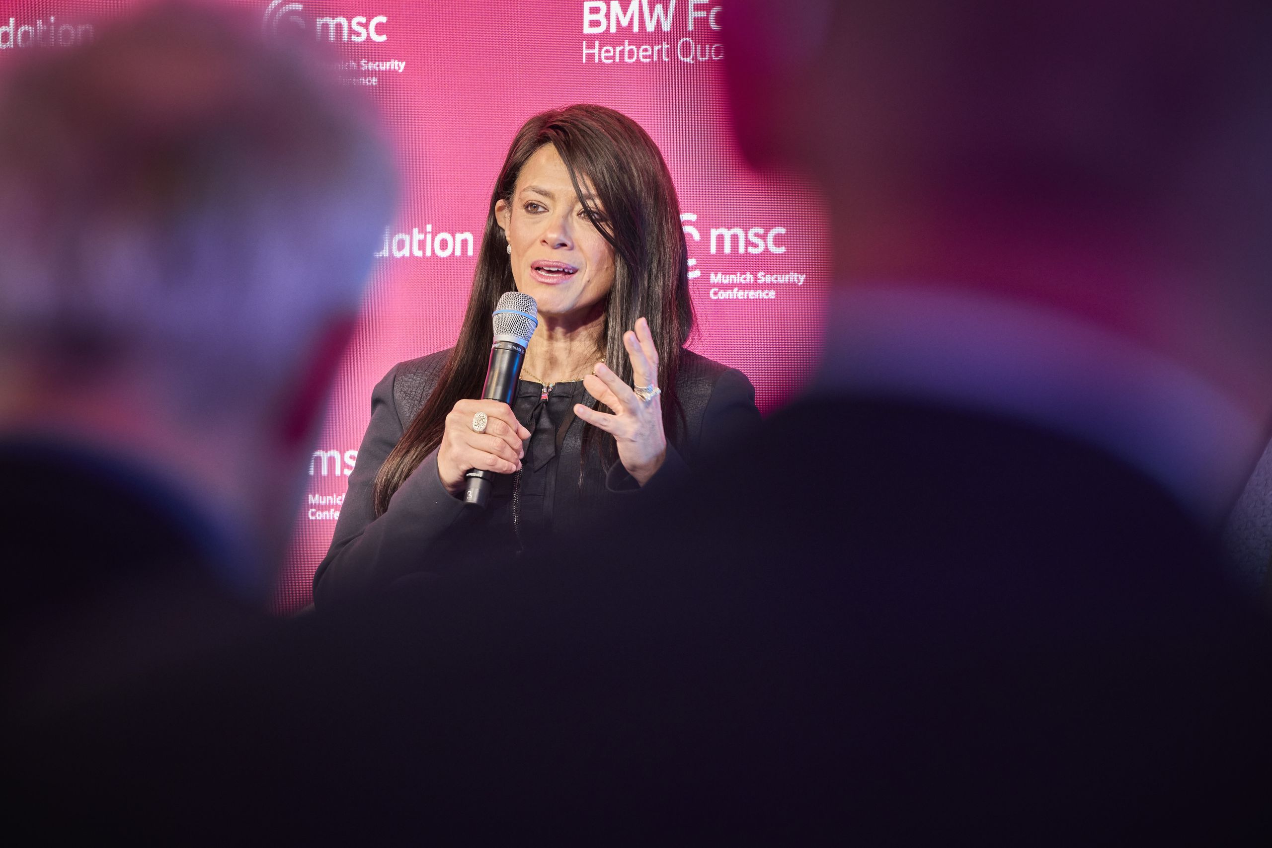 A woman holding a microphone speaks at an event with a red backdrop featuring logos, surrounded by blurred audience members.