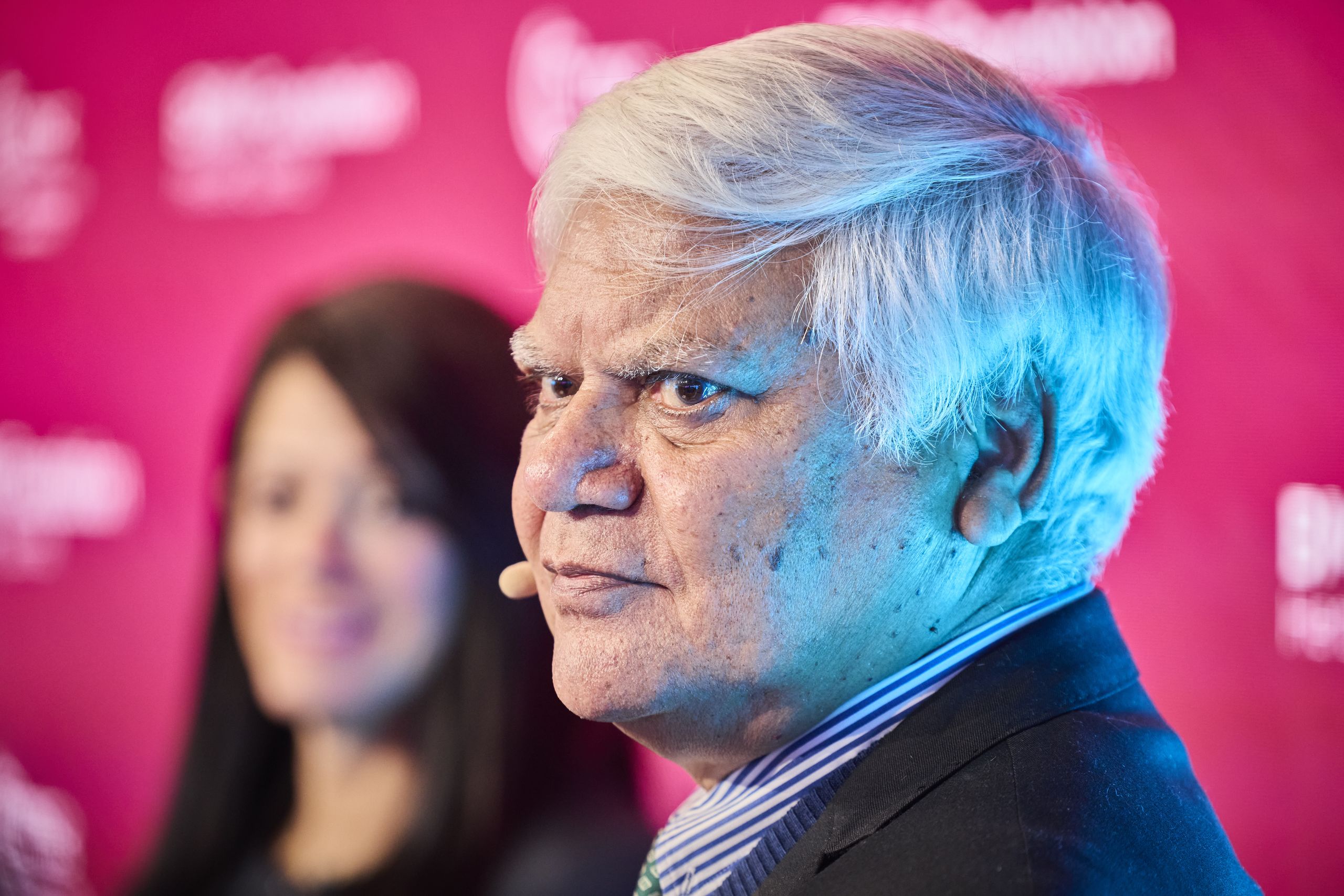 Elderly man with white hair and microphone headset, facing slightly right, with a woman in the blurred background, against a red backdrop.