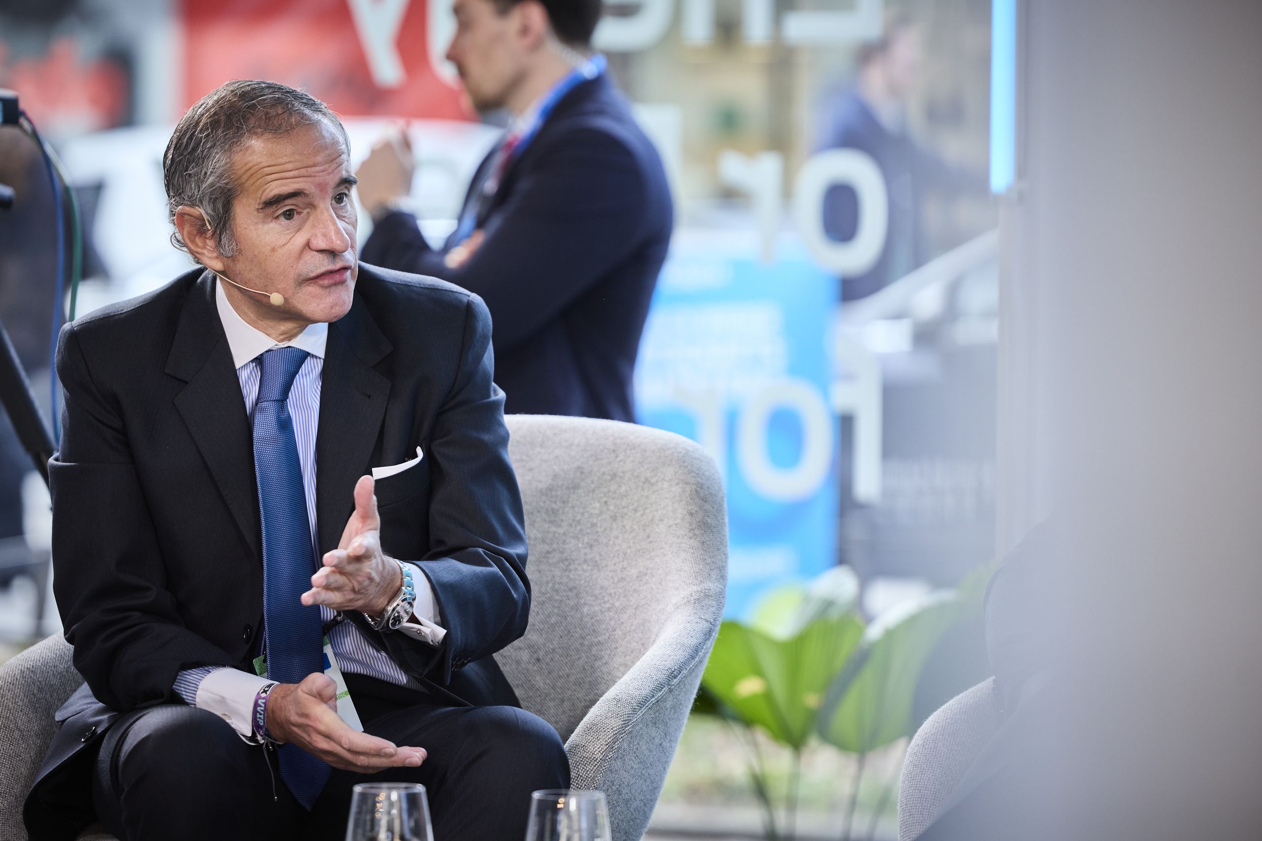 Man in a suit speaking during an interview, sitting on a gray chair. Blurred background with people and large windows.