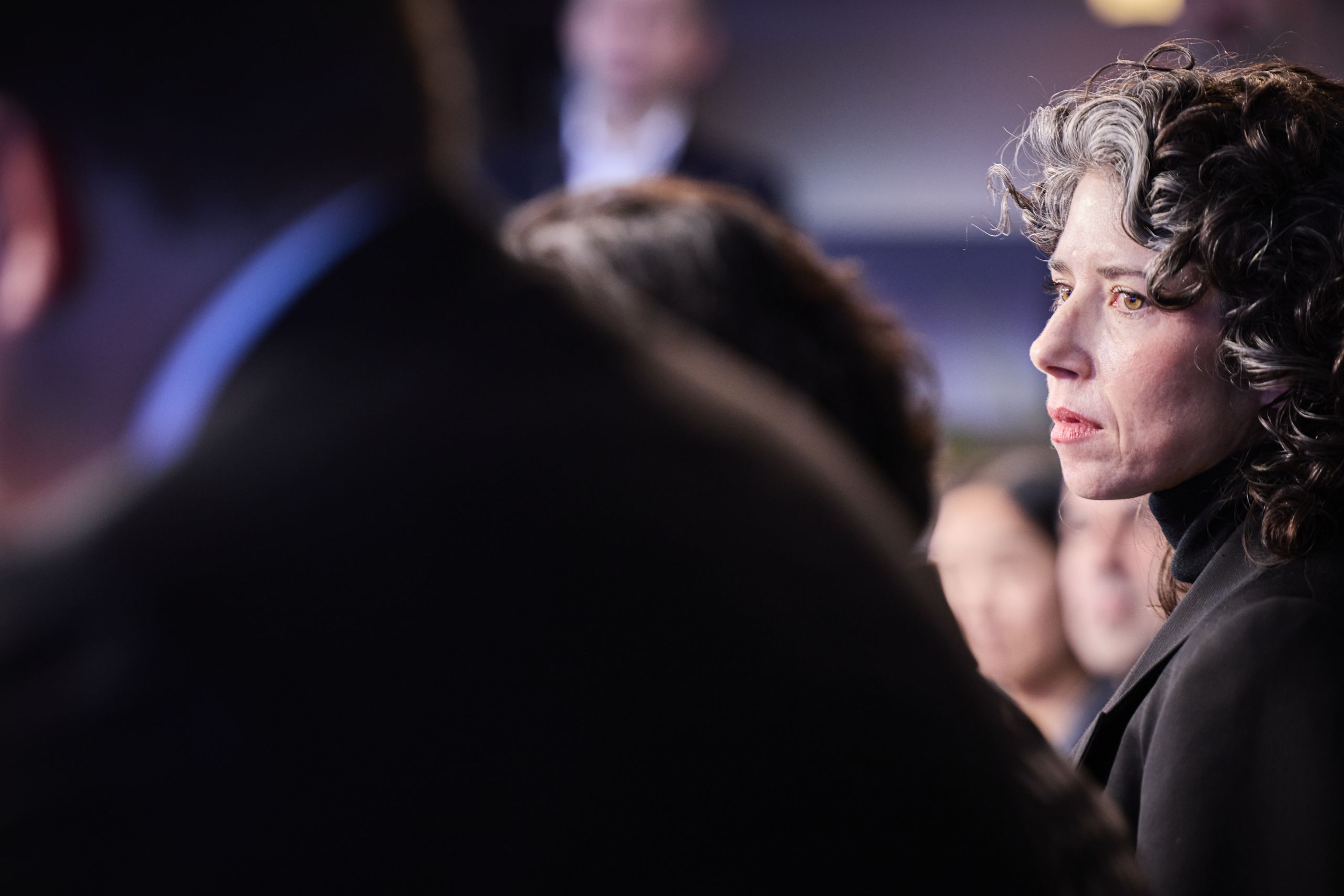 Woman with curly hair attentively listening in a crowd, with blurred figures in the foreground.