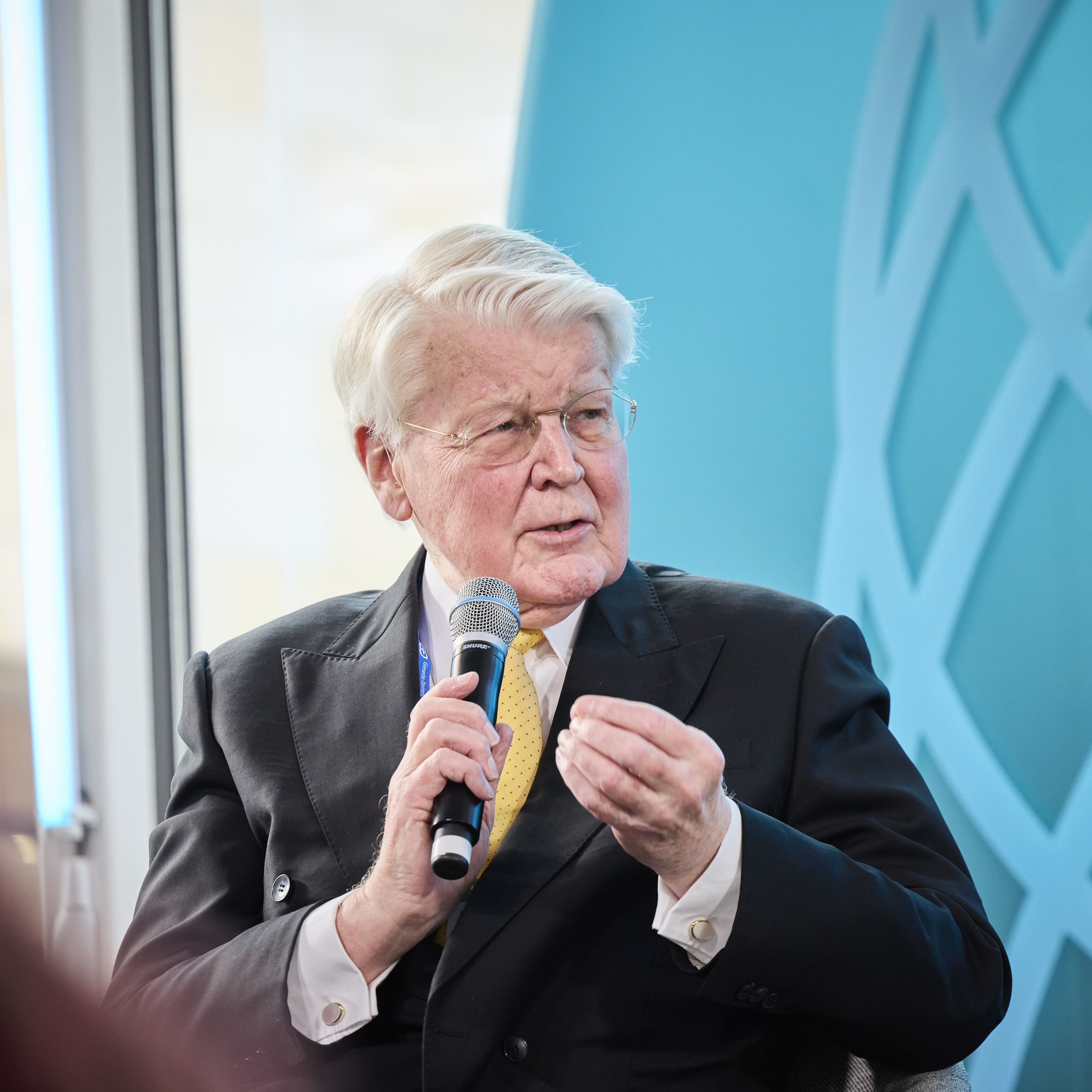 Elderly man in a suit speaking into a microphone at an event, with a blue backdrop and globe illustration.