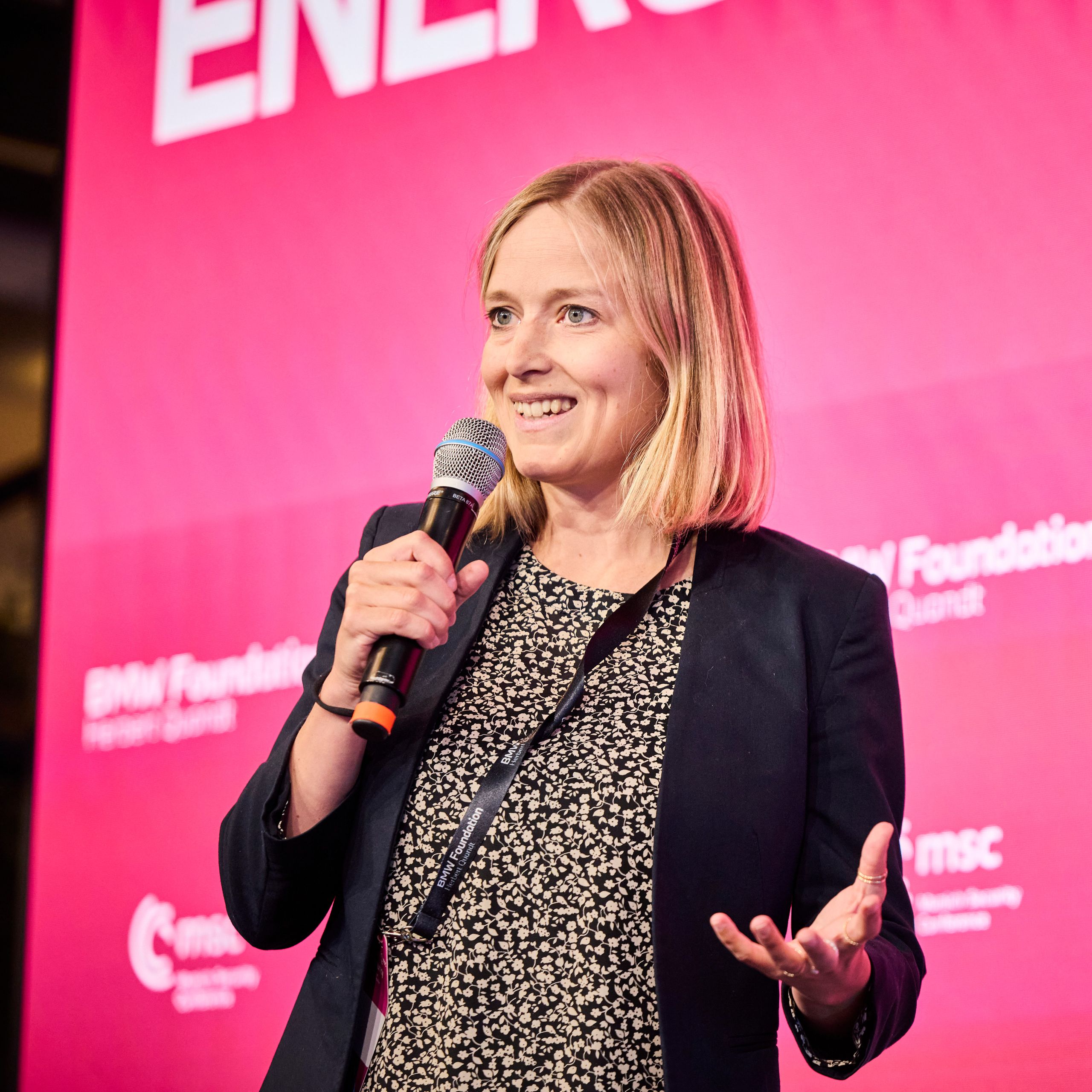 Woman speaking on stage with a microphone, standing in front of a red backdrop featuring logos and text for BMW Foundation Herbert Quandt.