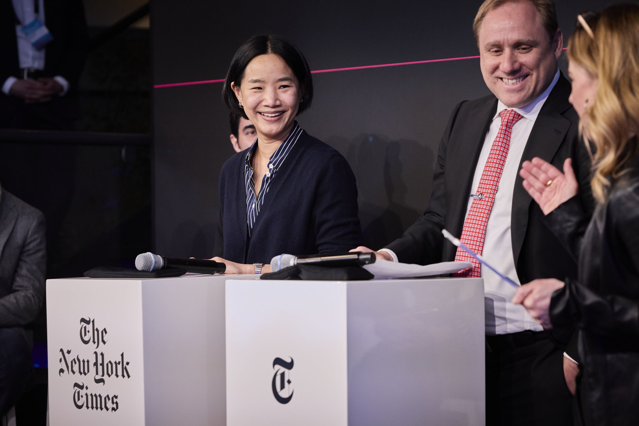 Three people in business attire stand and smile on a stage behind podiums with "The New York Times" logo. One woman is speaking animatedly.