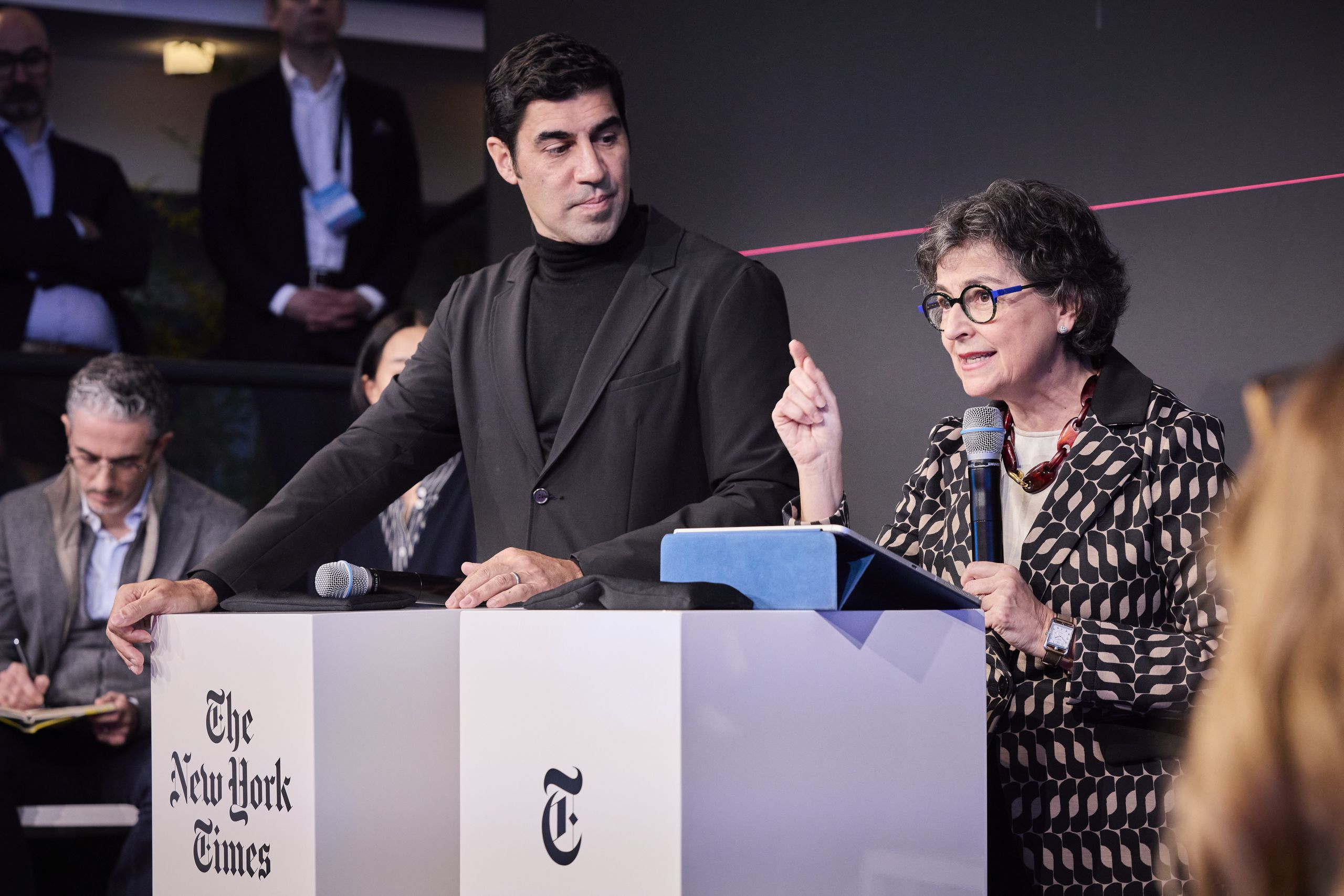 Two people stand at a podium with The New York Times logo. One speaks into a microphone, gesturing with a finger, while the other listens.