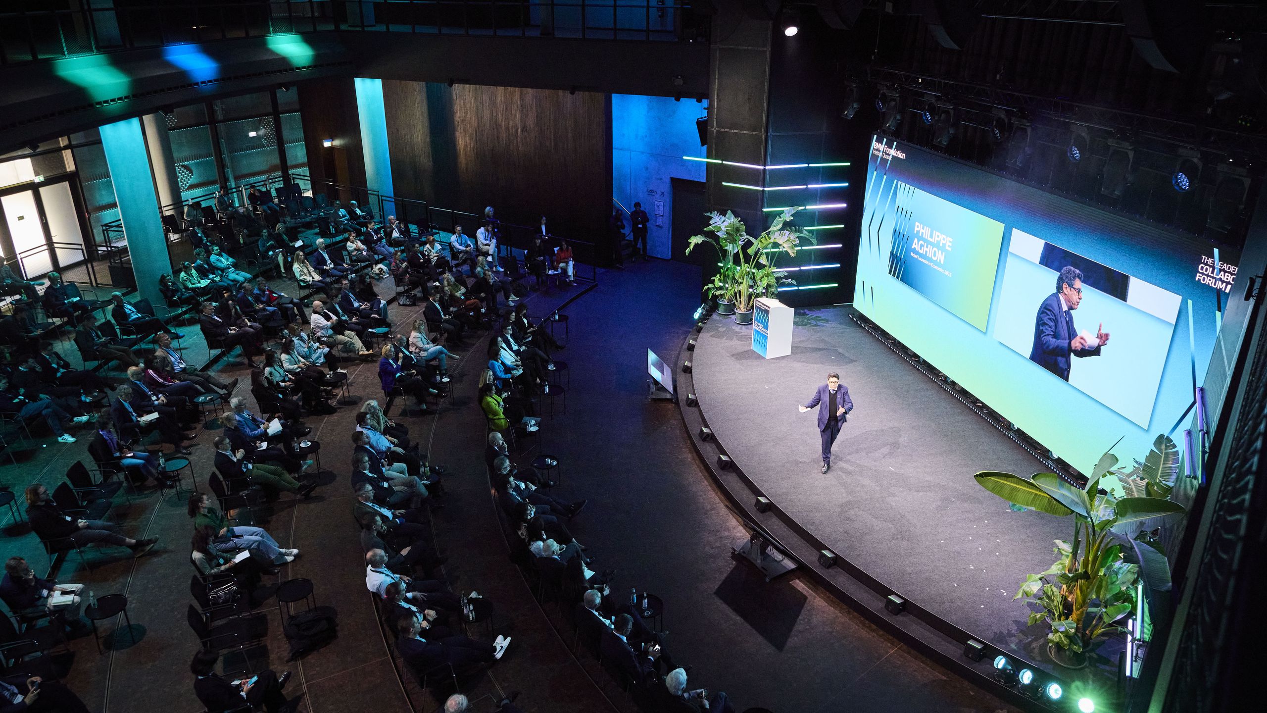 Philippe Aghion giving his keynote speech at The Leaders Collaboration Forum 2026 in Berlin. A speaker on stage presents to a large audience in a modern auditorium, with a big screen displaying visuals and green plants decorating the area.