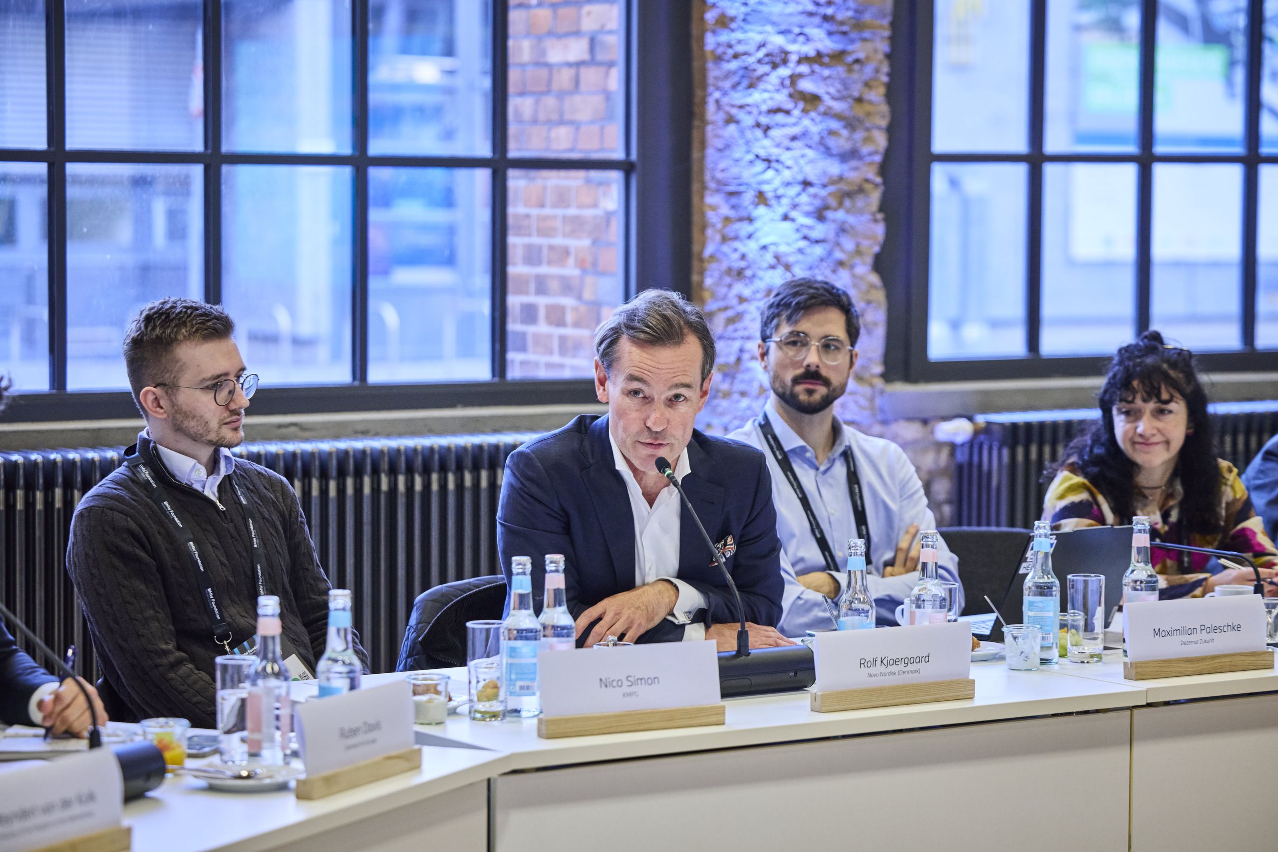 Rolf Kjærgaard, Special Advisor, Novo Nordisk Foundation. A man speaking at a conference table with several other people, bottles, and nameplates in front of a large window.
