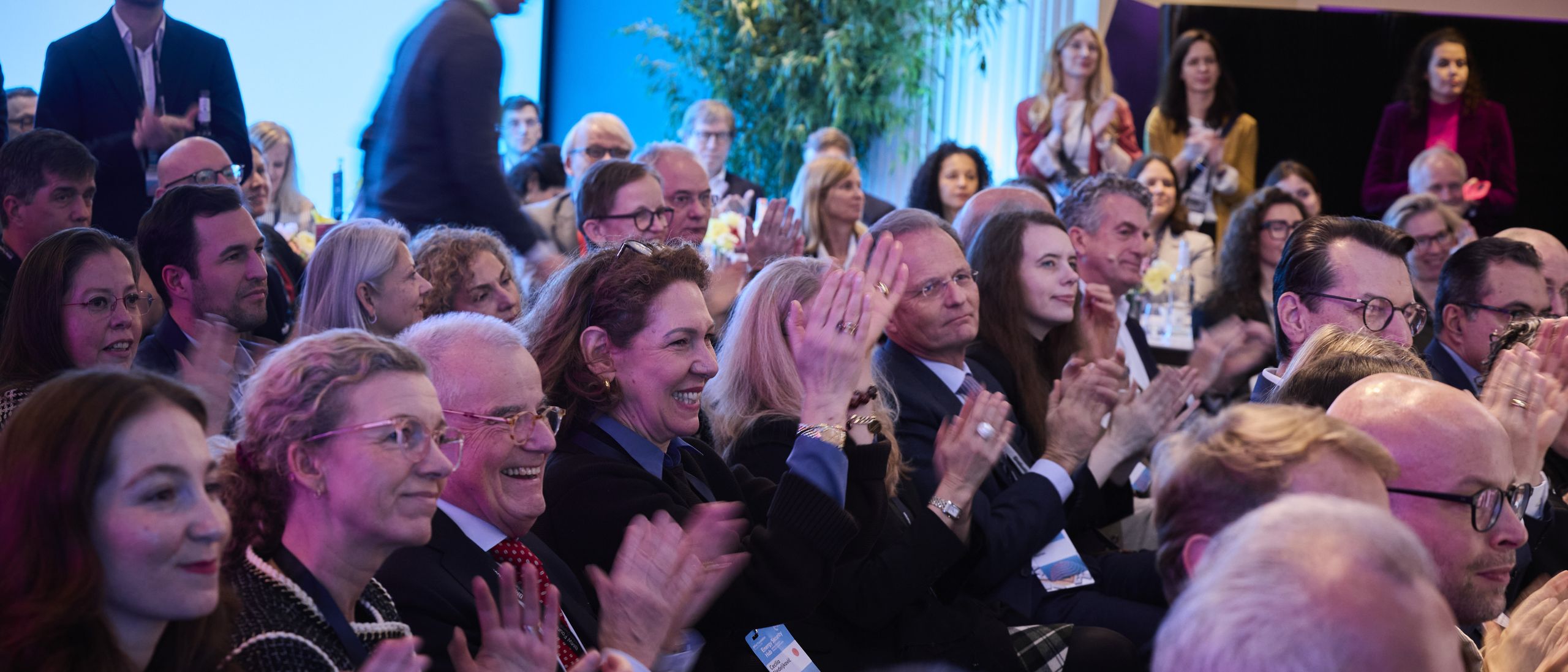 Audience clapping enthusiastically at an indoor event, with people of various ages and backgrounds seated closely together.