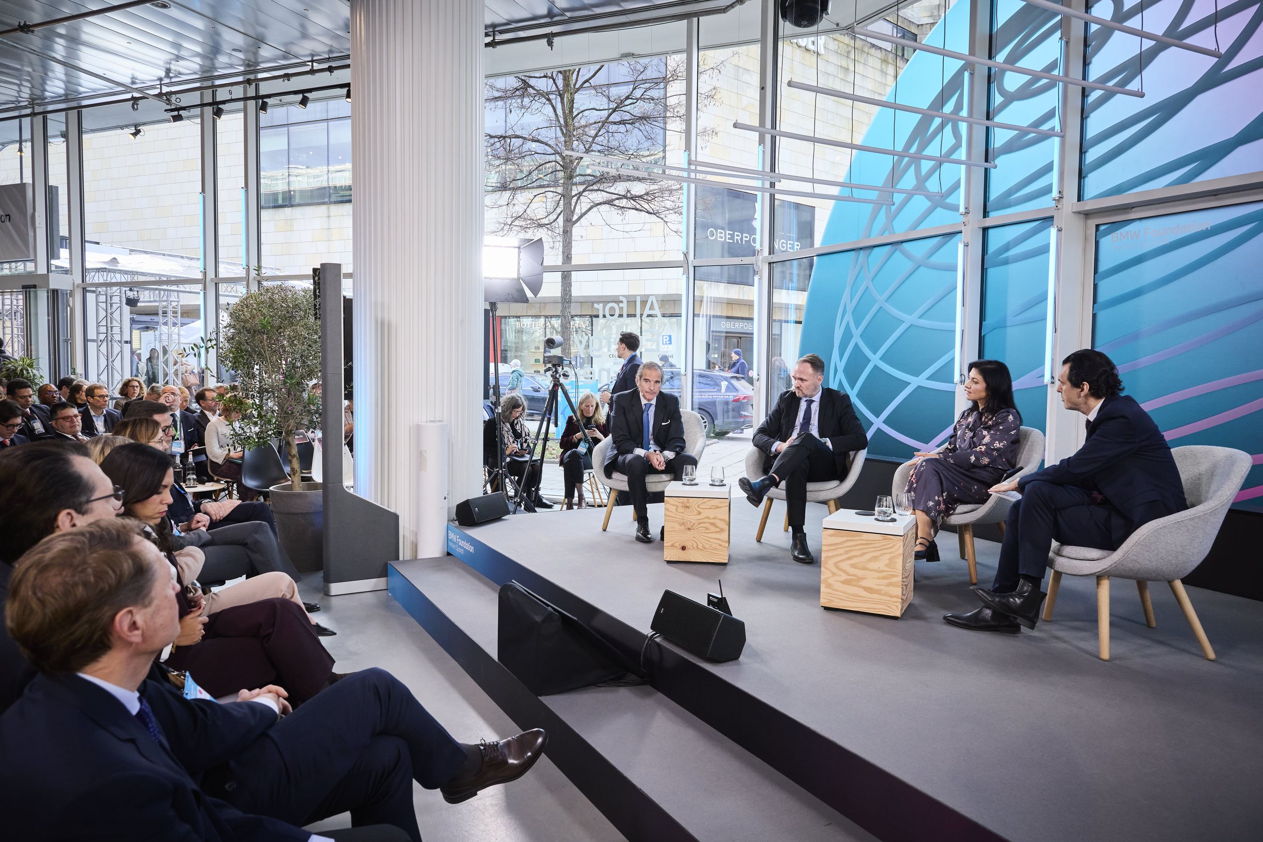 Panelists seated on stage in a modern conference room with an audience watching, large windows in the background.