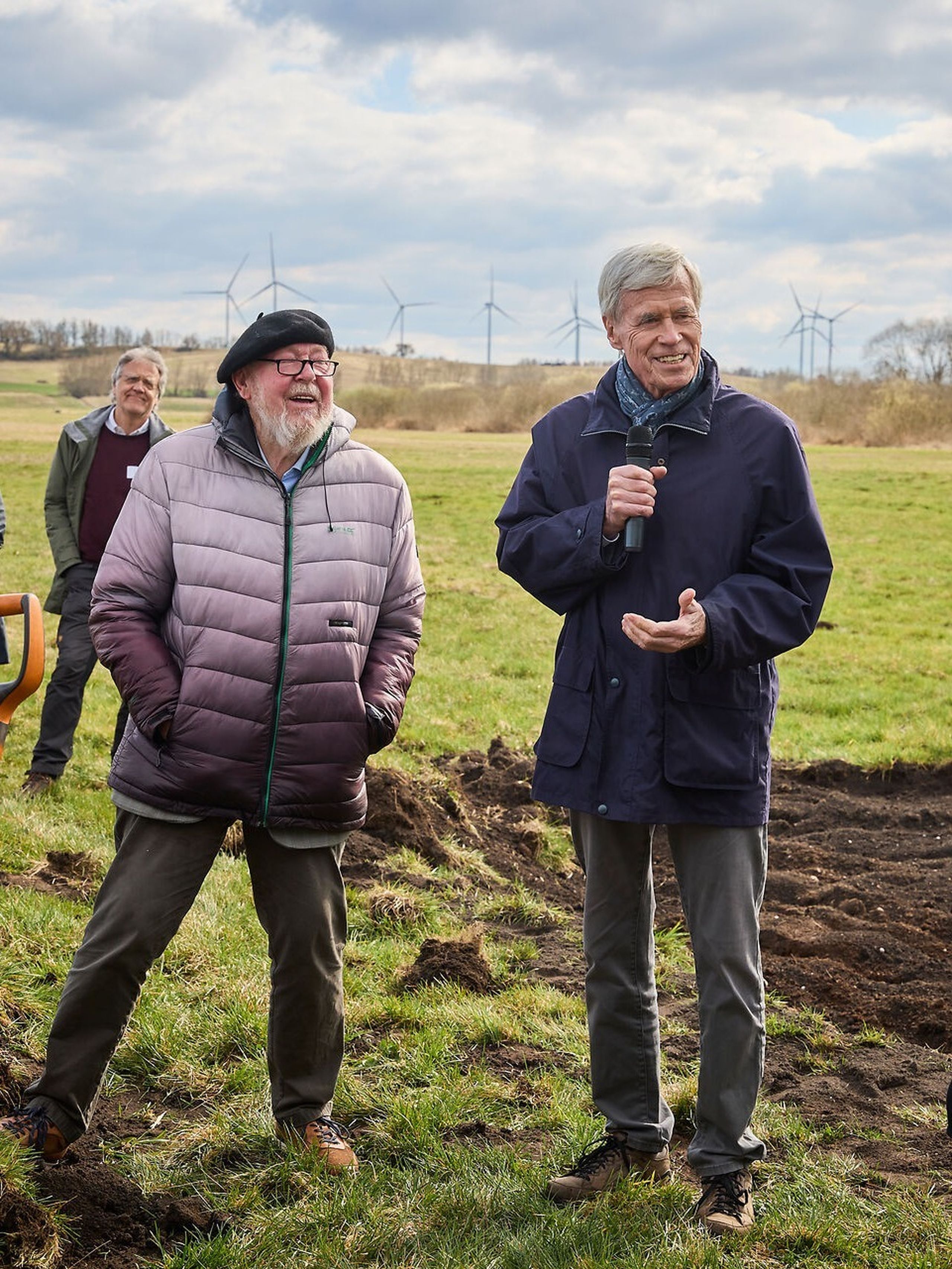 Michael Succow (left) and Michael Otto during the project launch. Photo: Marc Beckmann Two men stand in a grassy field, one holding a microphone. Wind turbines are visible in the background under a cloudy sky.