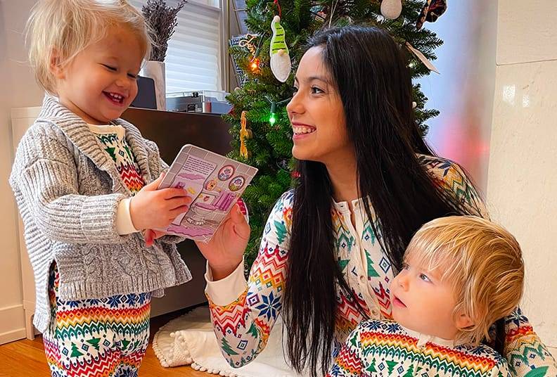 Smiling au pair and two happy toddlers in matching colorful holiday pajamas by a decorated Christmas tree.