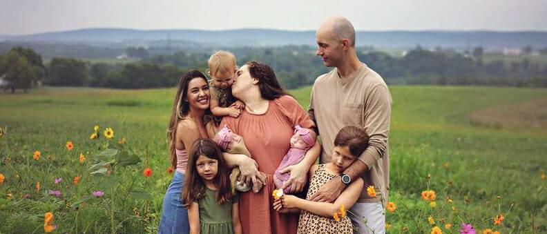 A family of eight, including parents, three children, two infants, and their au pair, stands in a colorful flower field under a cloudy sky.