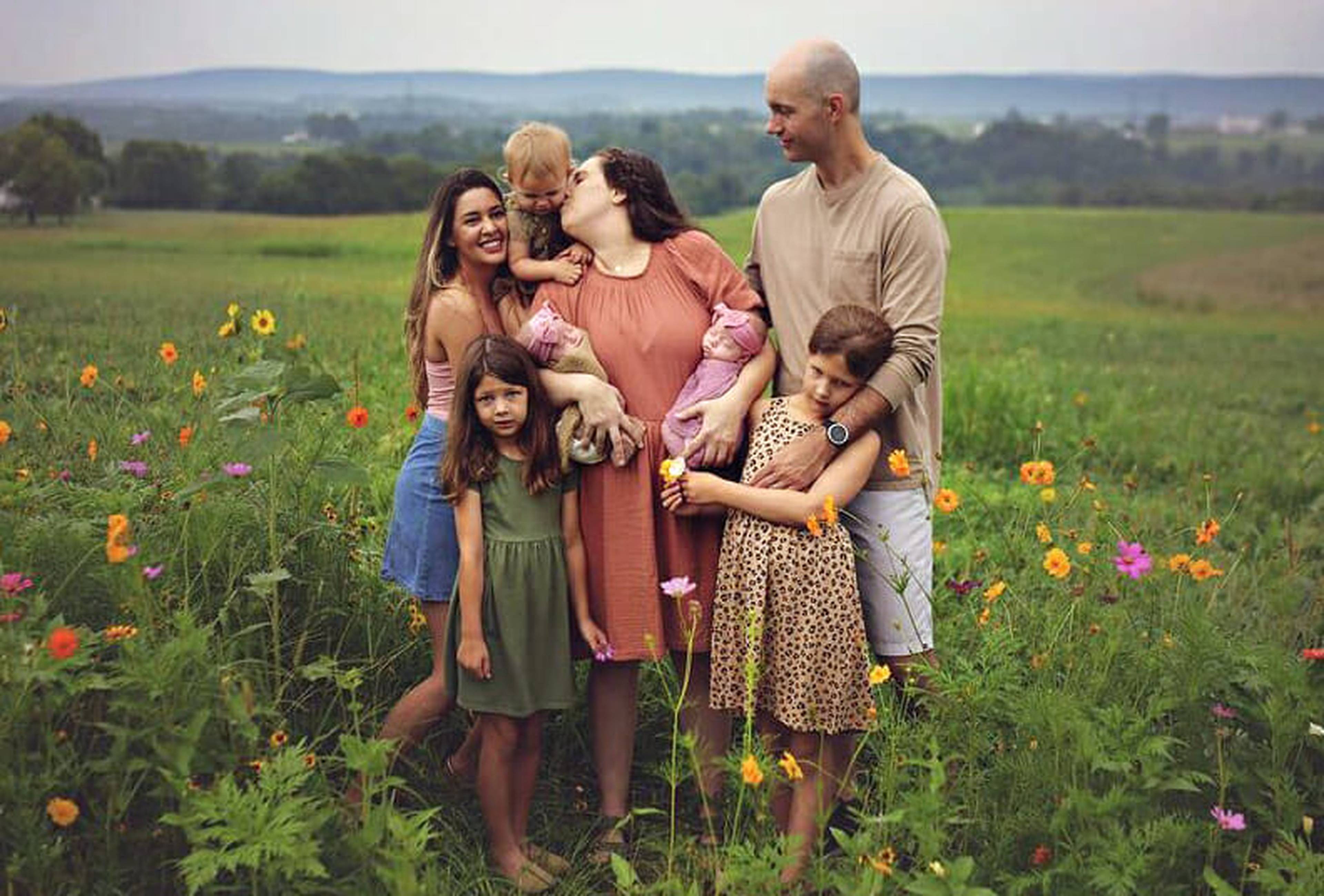 A family of five with their au pair stands in a colorful flower field, with a woman kissing a baby and children looking at the camera.