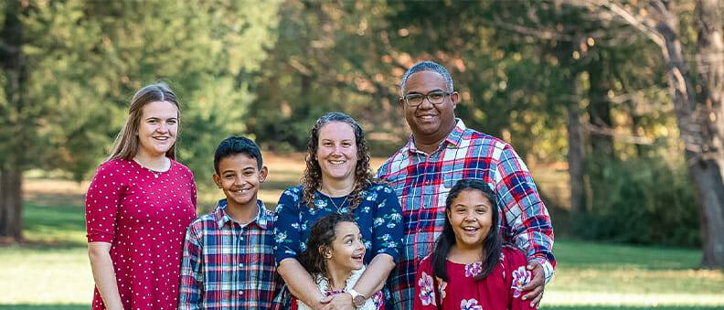 A multi-racial host family with two parents, three kids, and their au pair pose outdoors on grass, wearing coordinated red and plaid outfits, with trees in the background.