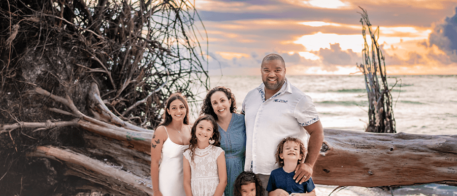 Family of six -- a tall Black man, a white woman, their three children, and their Italian au pair - posing on a sandy beach at sunset, with driftwood and ocean waves in the background.