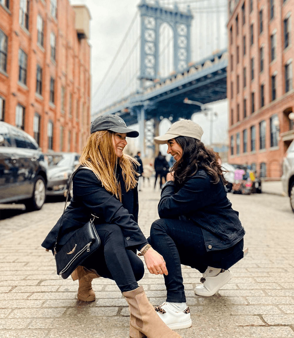Two women in casual attire crouch and smile at each other on a cobblestone street, with a suspension bridge in the background.