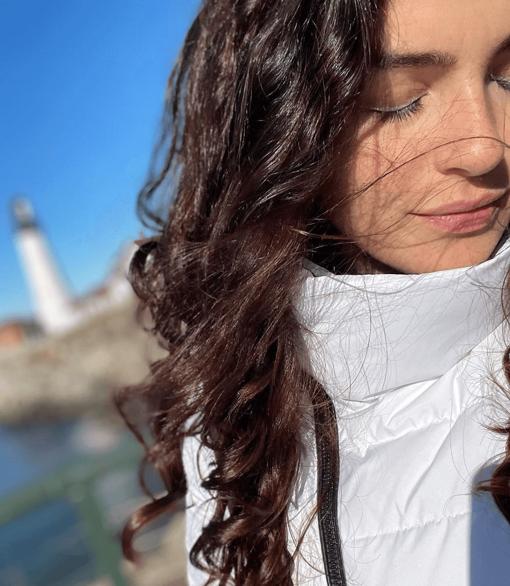 Woman with long curly hair wearing a white jacket, eyes closed, with a blurred lighthouse and blue sky in the background.