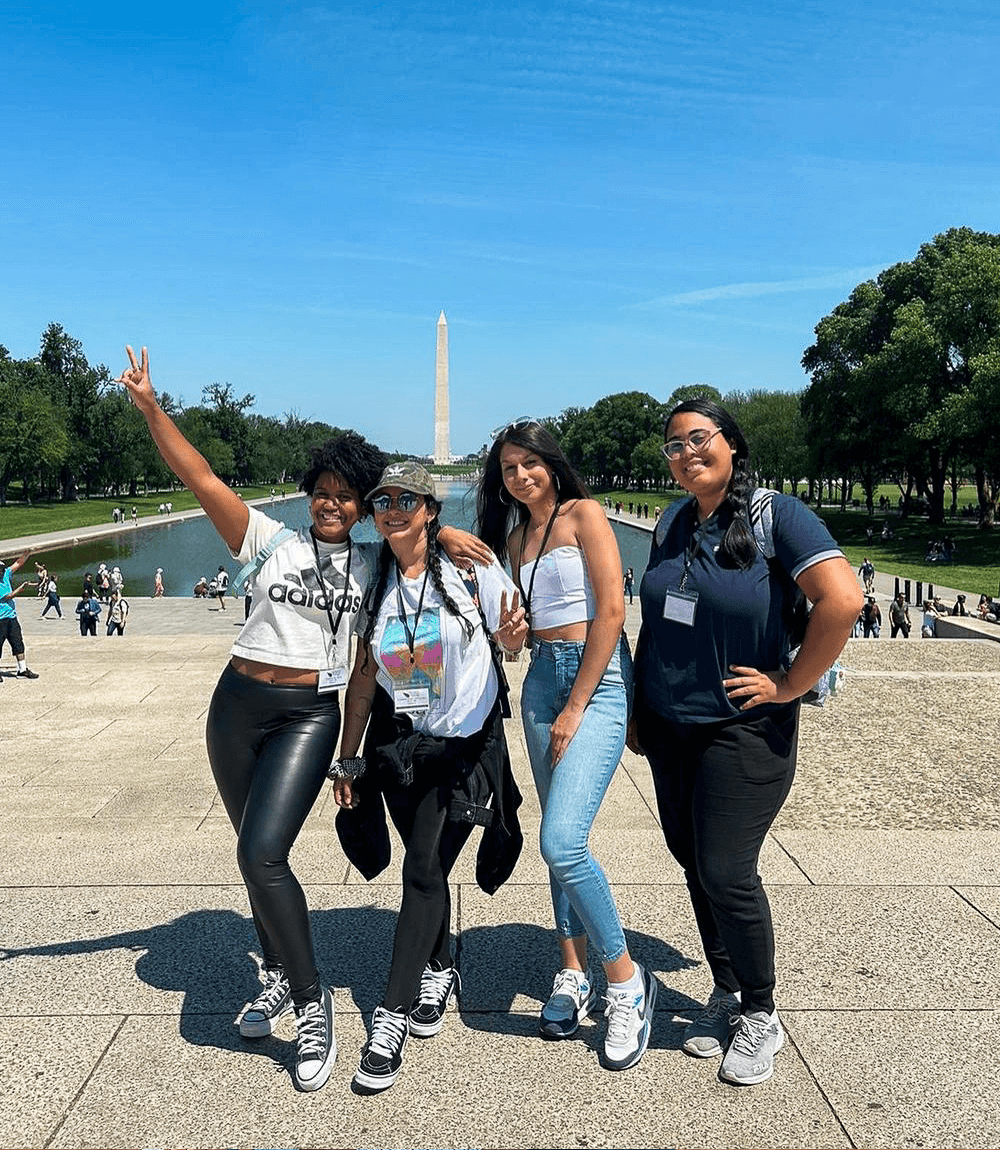 Four people pose happily in front of the Washington Monument under a clear blue sky, with one making a peace sign.