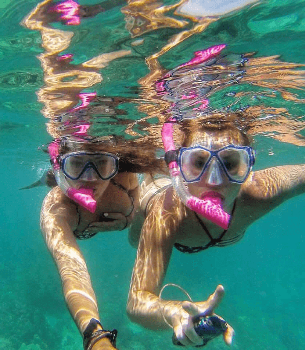 Two people snorkeling underwater, wearing masks and snorkels, with sunlight creating patterns on their skin and the water’s surface.