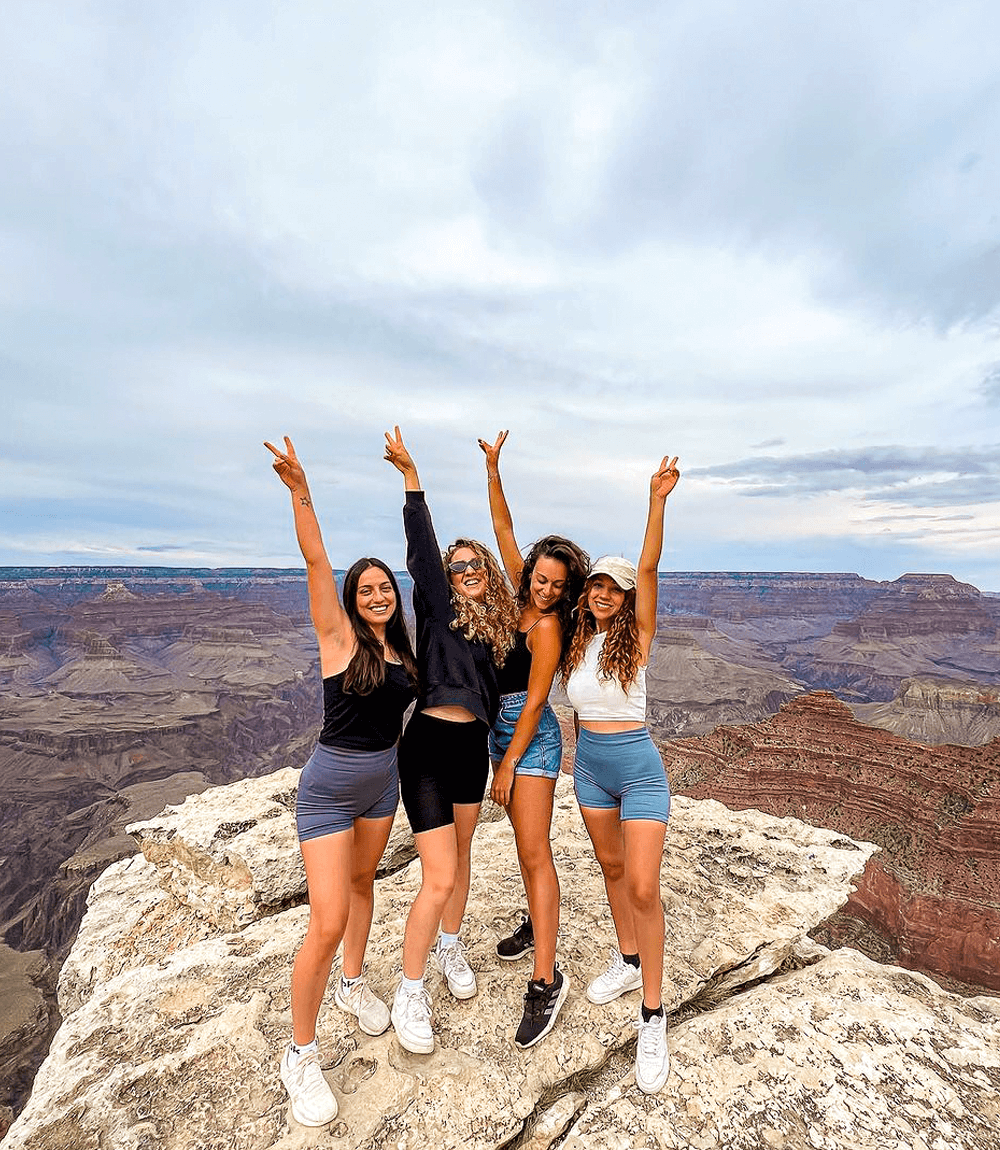 Four women smile and raise their arms in celebration while standing on a rocky cliff with the Grand Canyon as a breathtaking backdrop.