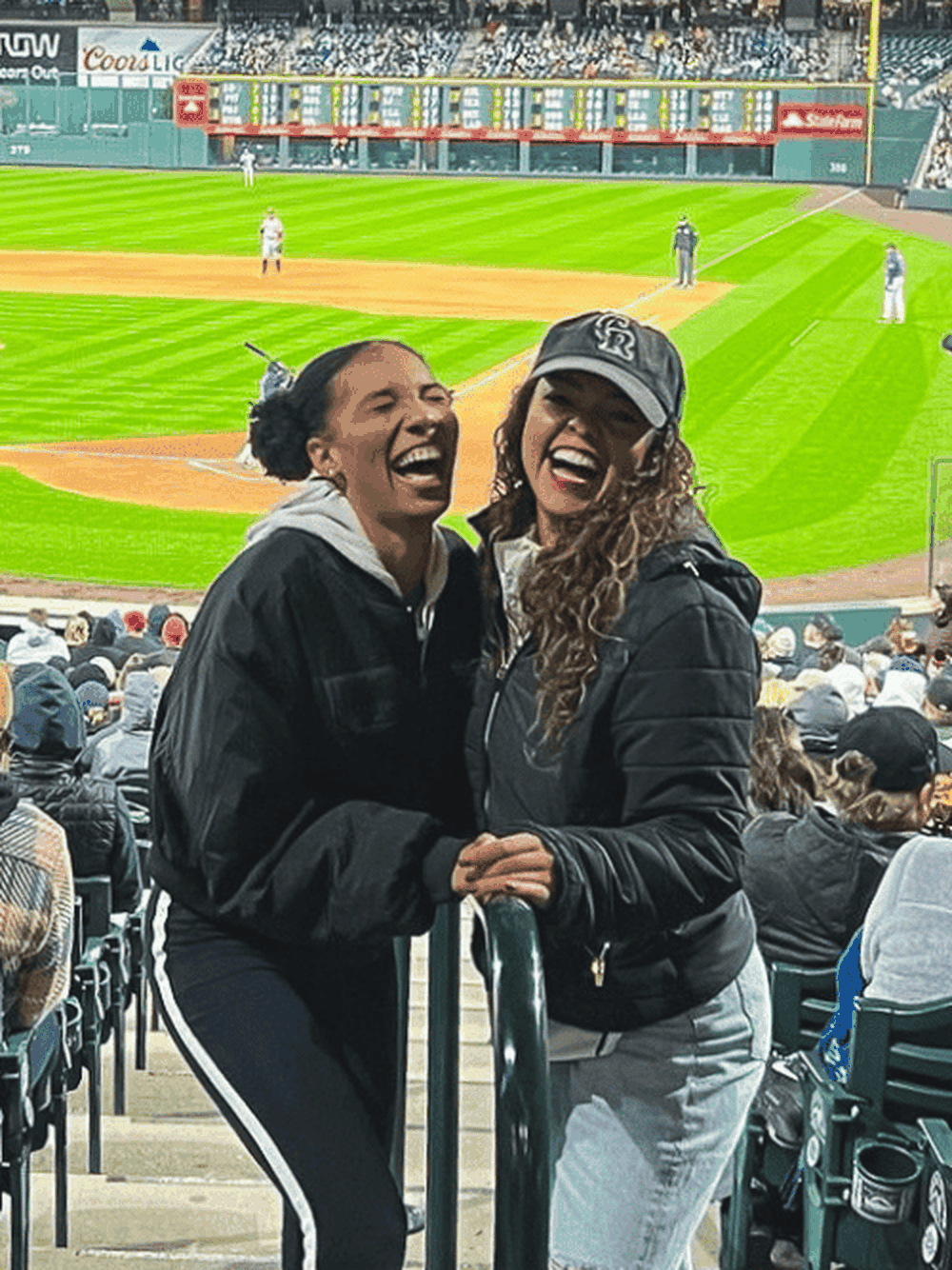 Two women laughing and posing for a photo in the stands at a baseball game, with the field and audience in the background.