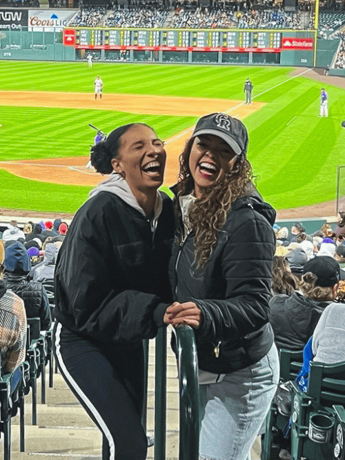 Two women laughing and posing for a photo in the stands at a baseball game, with the field and audience in the background.