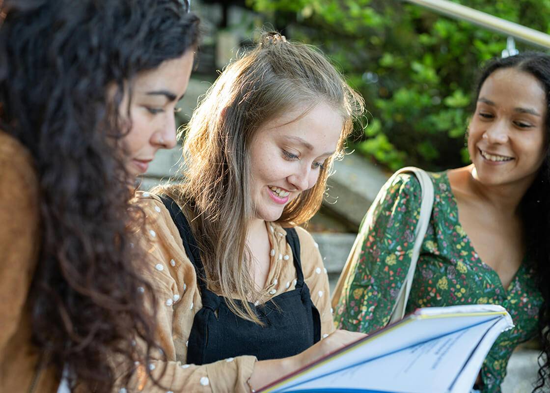 Three women sitting on outdoor steps, looking at a book together.