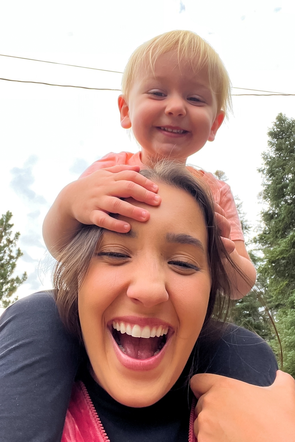 A woman joyfully carrying a smiling toddler on her shoulders outdoors, surrounded by trees. Both are laughing and facing the camera.