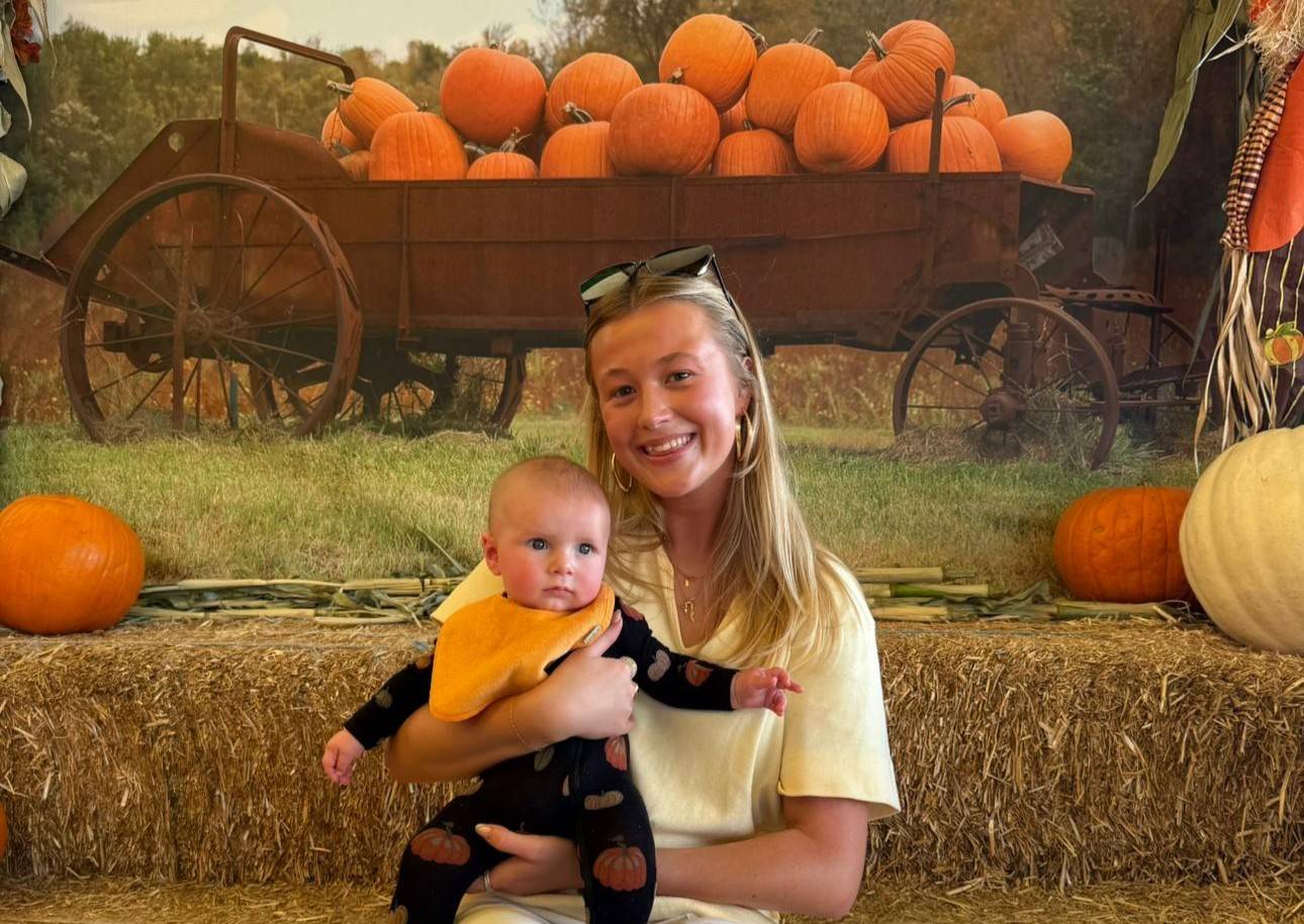 Smiling woman holds a baby wearing an orange bib, sitting on hay bales with pumpkins and an antique cart in the background.