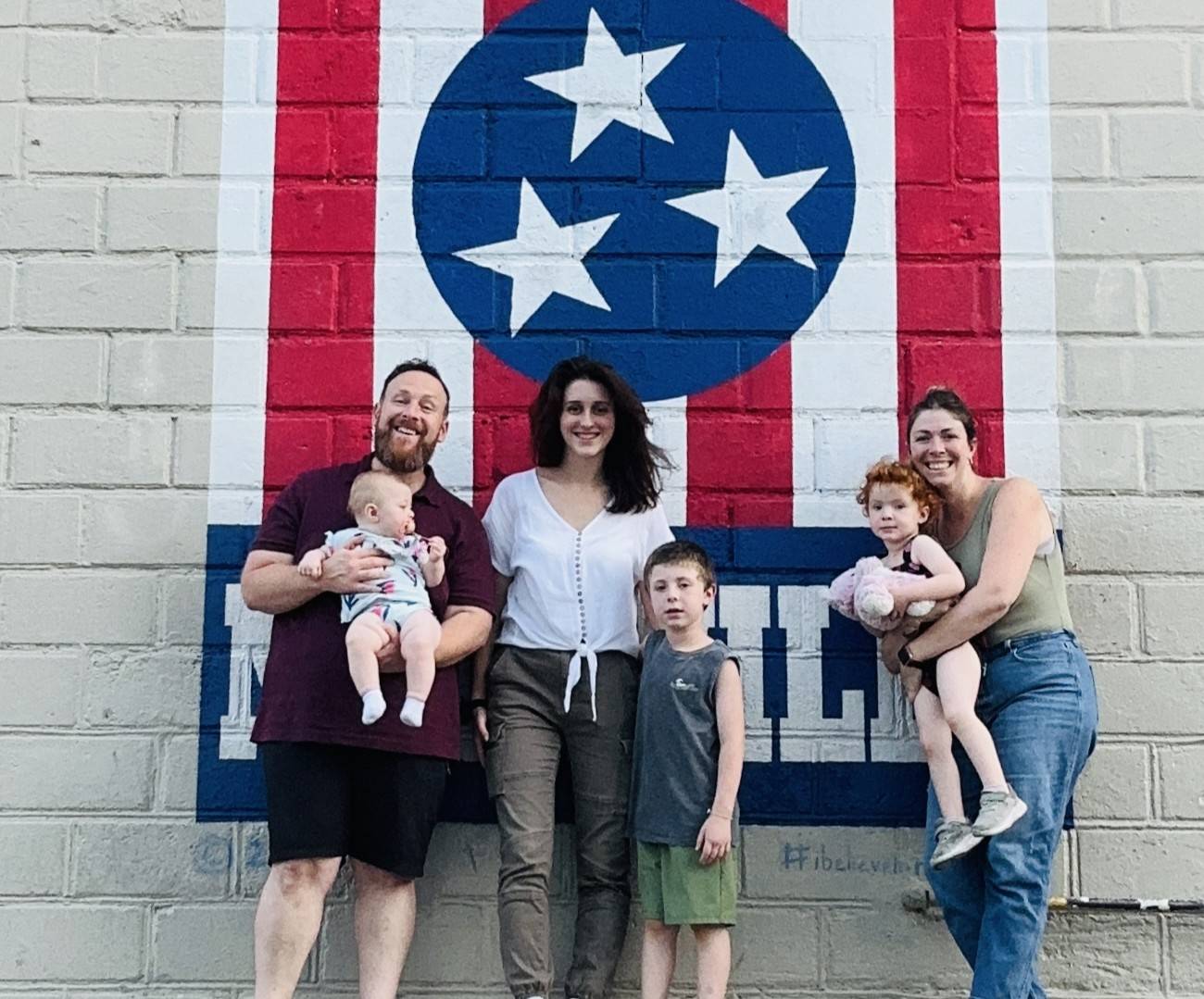 A group of adults and children stand smiling in front of a mural featuring three stars inside a circle, painted on a brick wall.