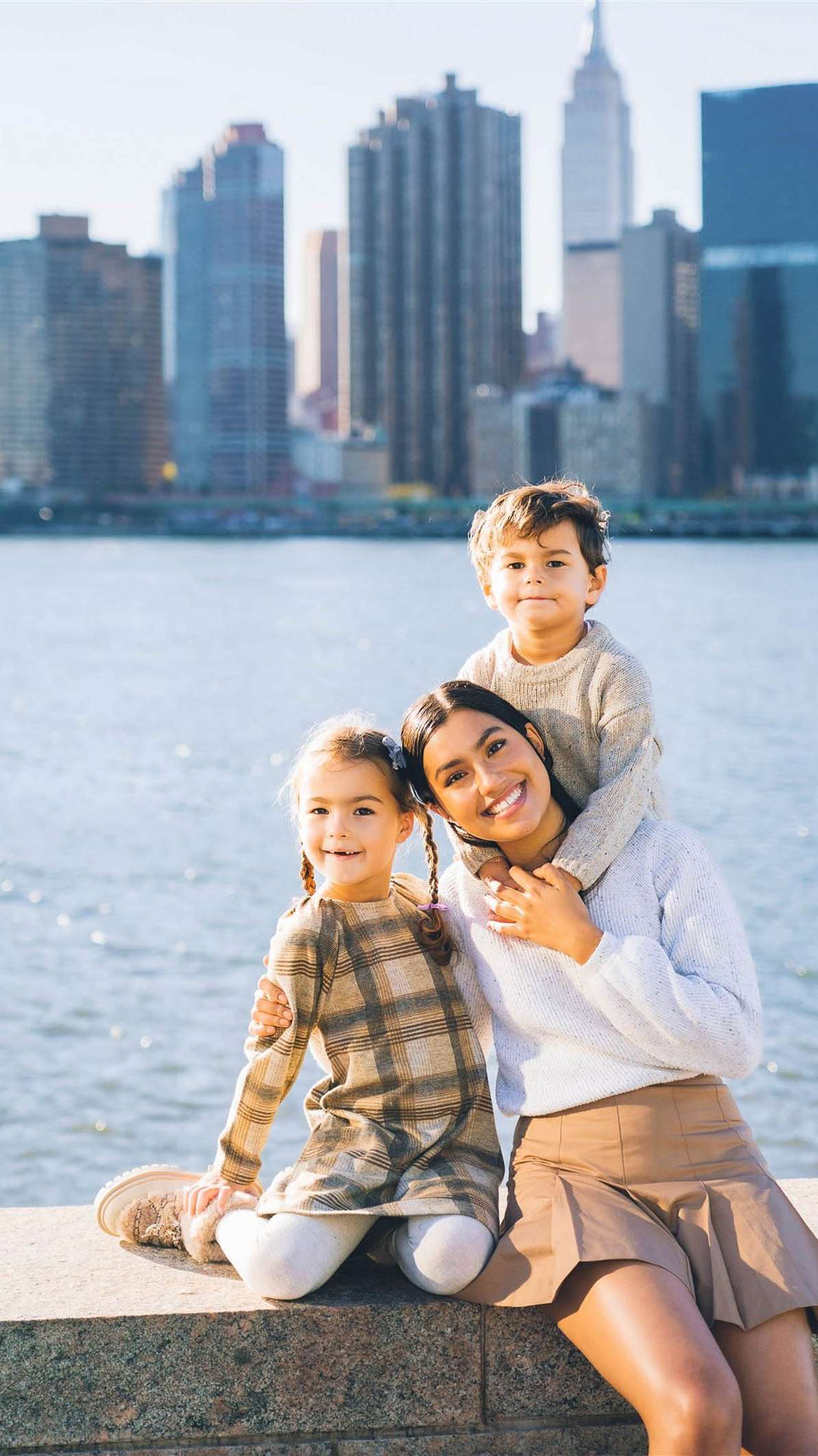 Woman with two children smiling by a waterfront, city skyline in the background.