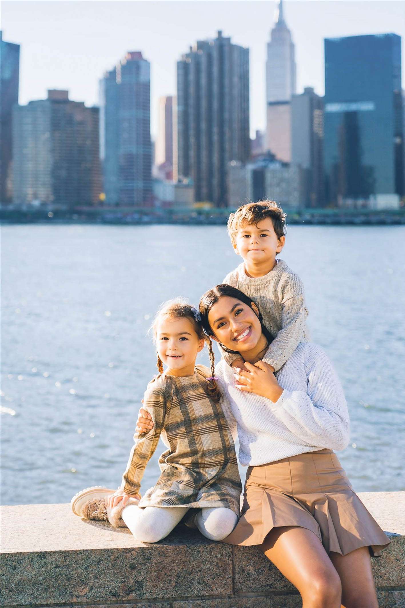 Woman with two children smiling by a waterfront, city skyline in the background.