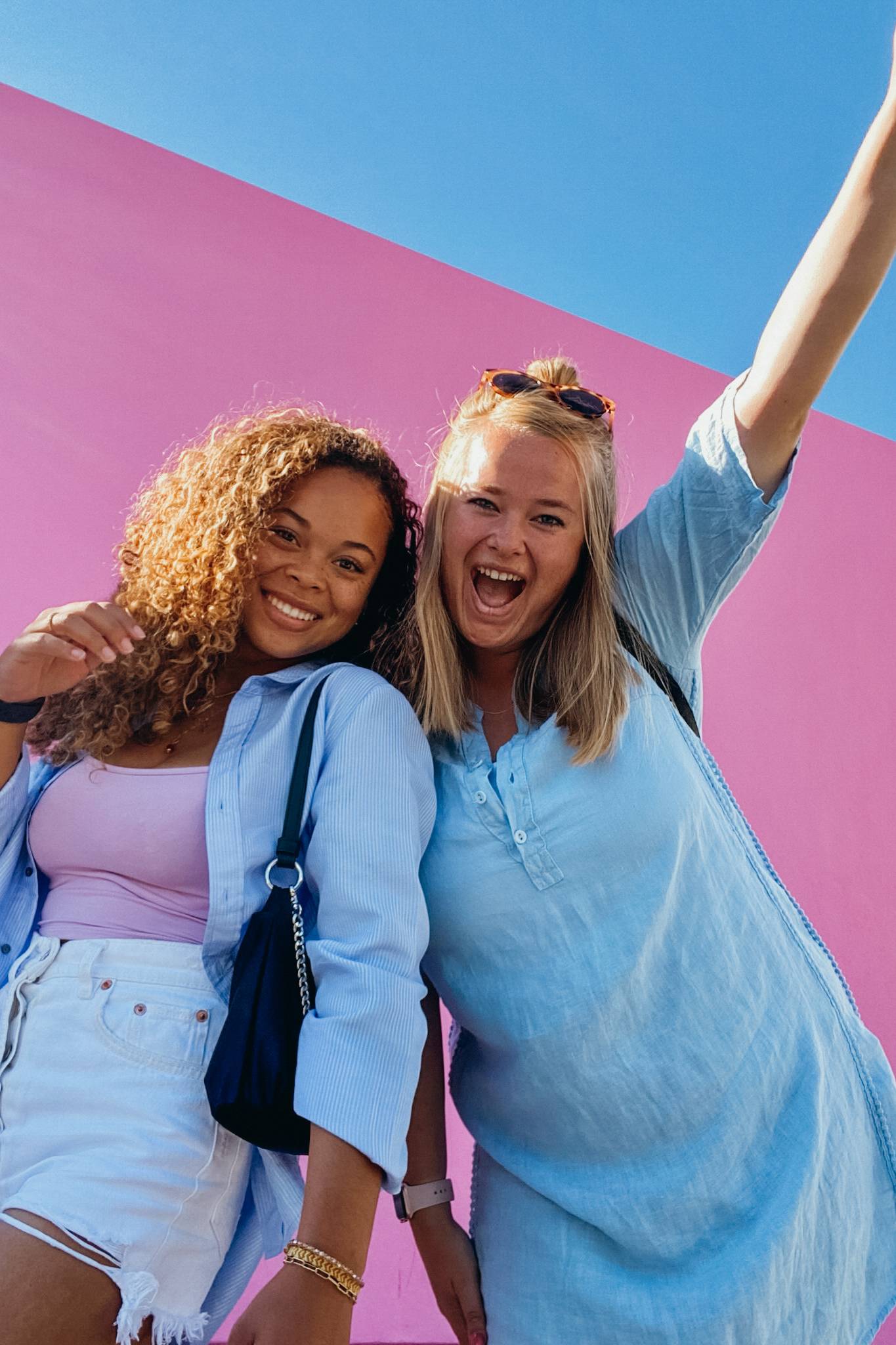 Two women smiling in front of a pink wall, one with a raised arm. They're wearing casual blue outfits against a clear blue sky.