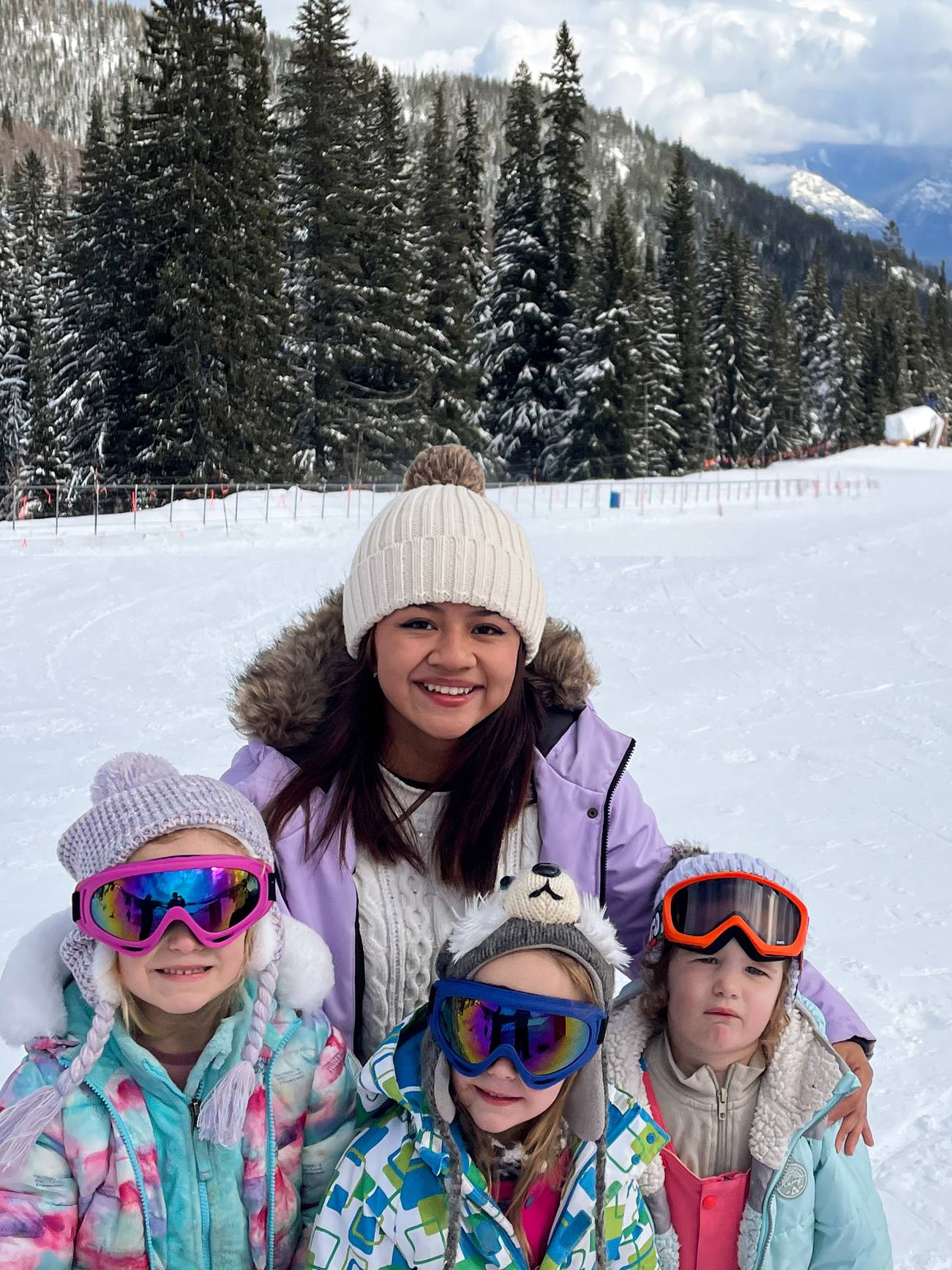A woman and three children wearing winter clothing and ski goggles pose on a snowy mountain slope with pine trees in the background.