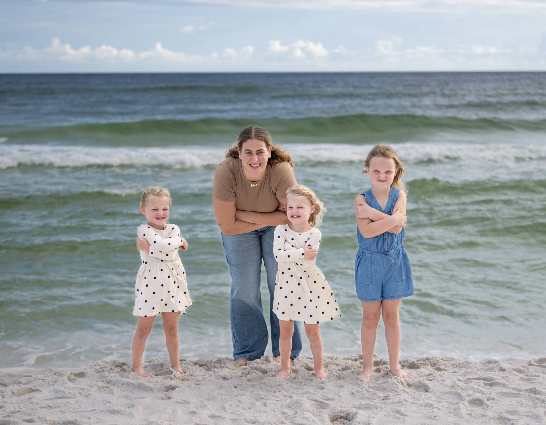 A woman and three young girls stand on a sandy beach with waves in the background, all smiling with arms crossed.