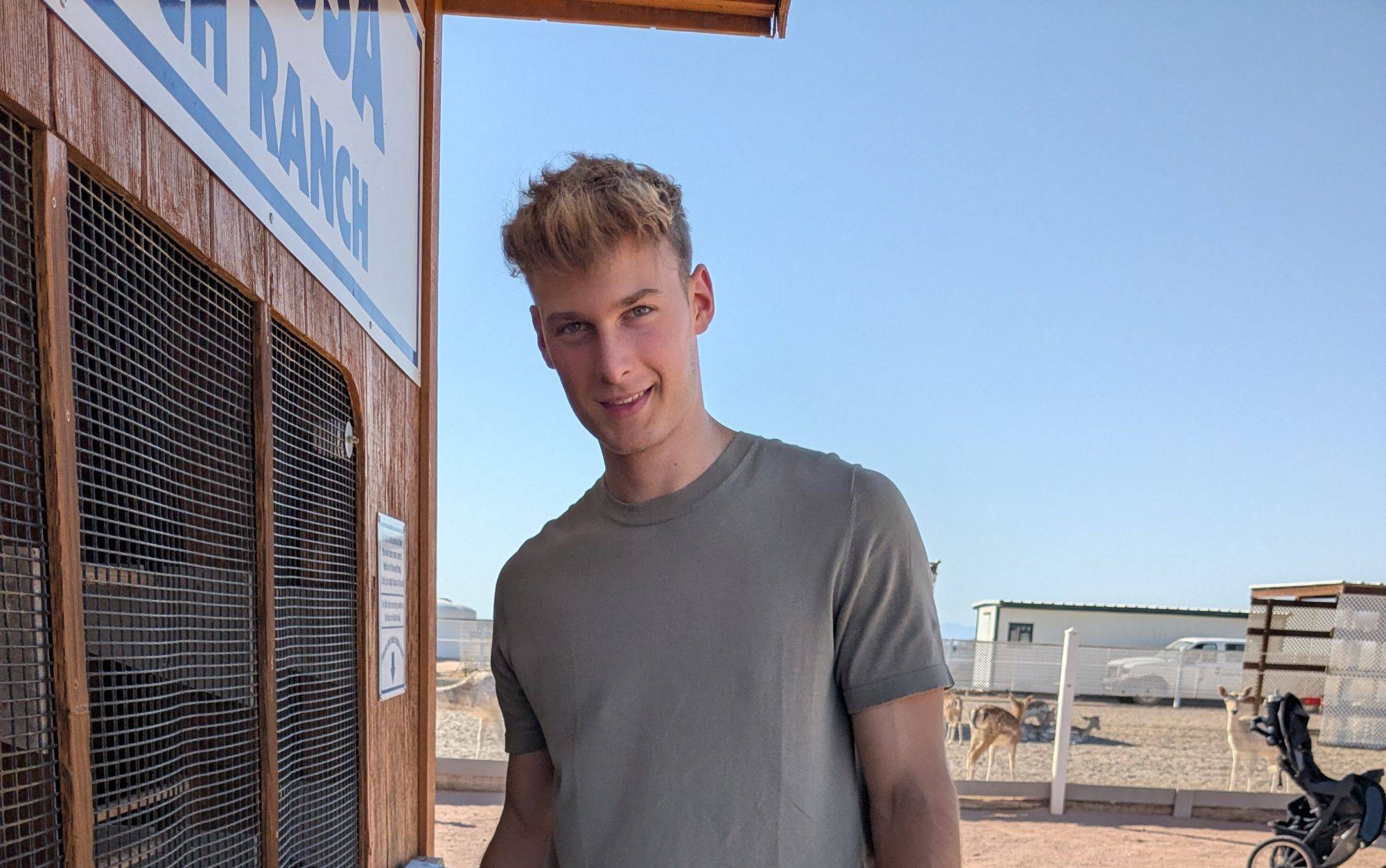 A person with short blond hair stands outside a wooden structure on a sunny day, with a fenced area and a small building in the background.
