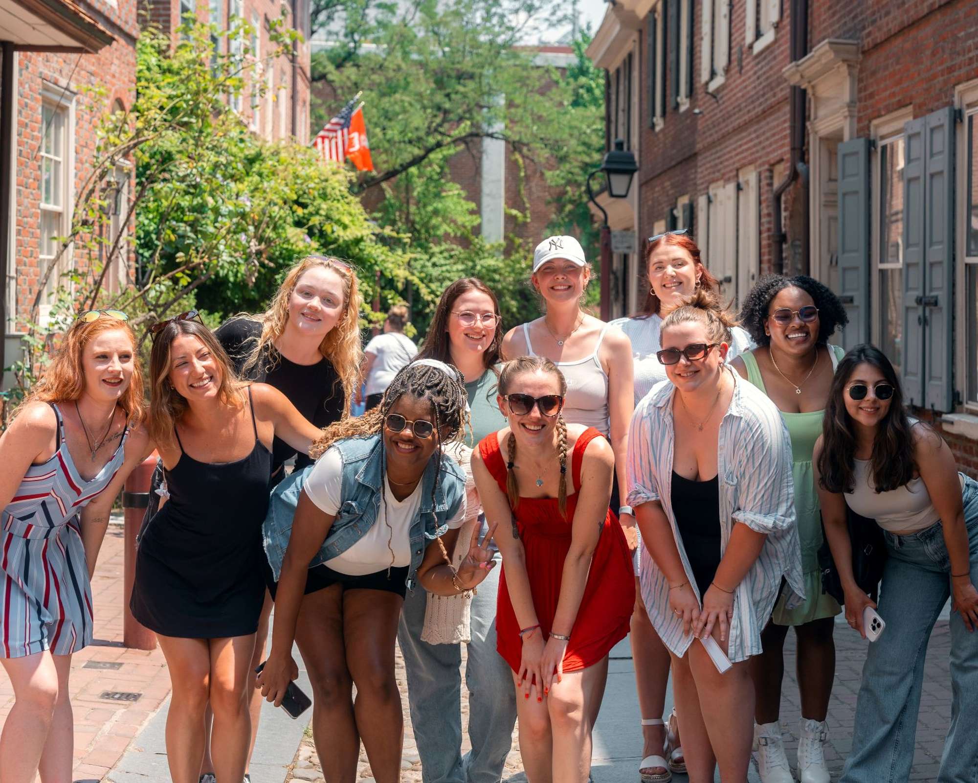 A group of smiling friends poses together on a sunny brick-lined street, with trees and historic buildings in the background.