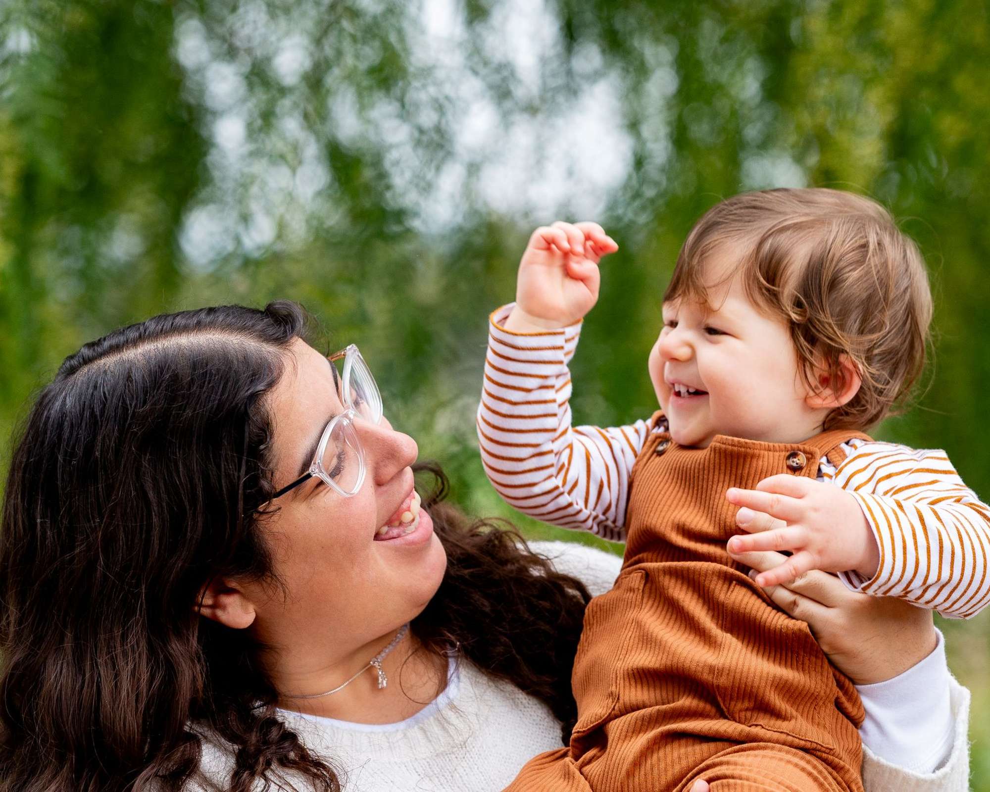 A joyful adult holding a smiling child in brown overalls outdoors, surrounded by greenery. Both are looking at each other happily.