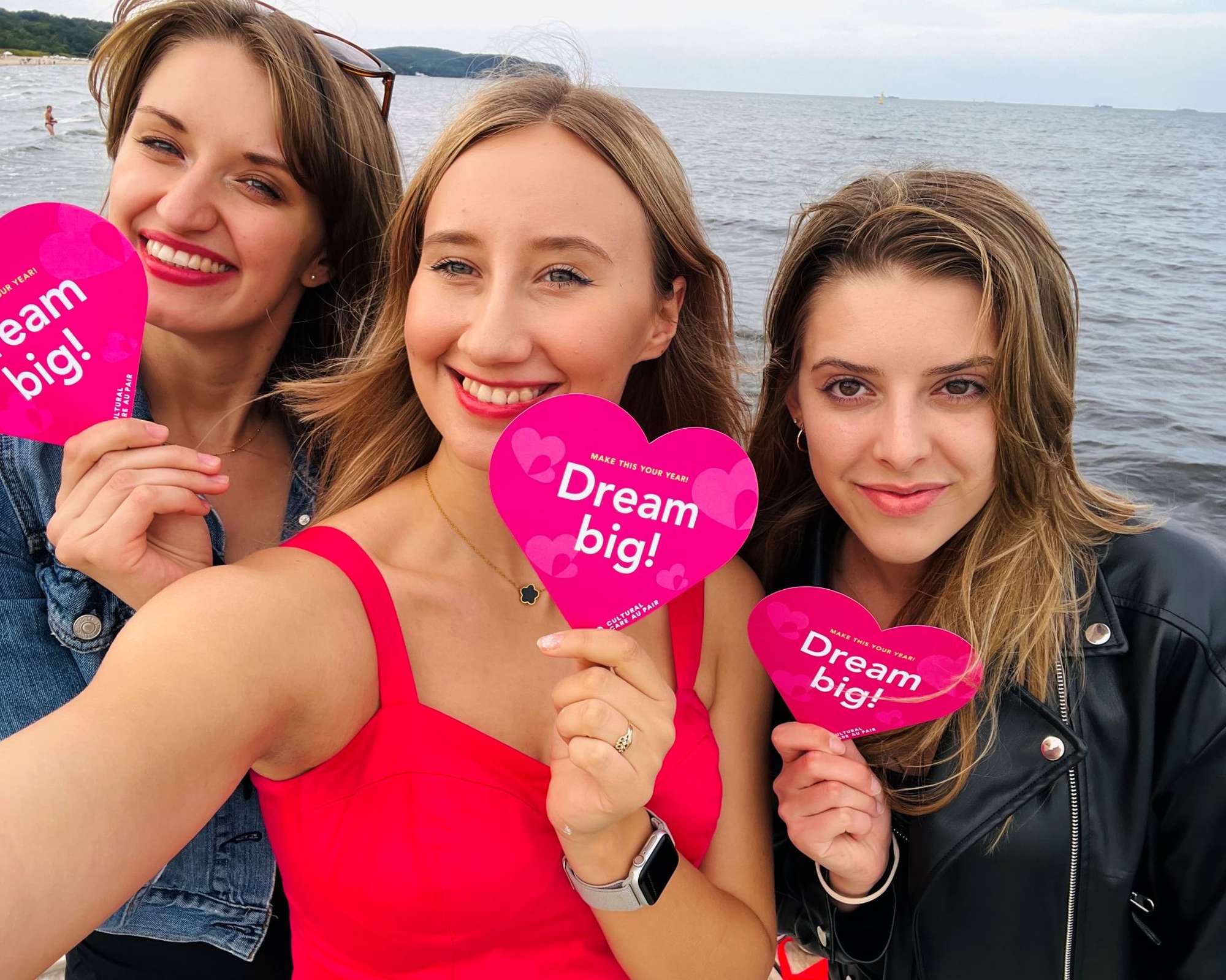Three smiling women at the beach hold pink heart-shaped signs reading "Dream big!" on a cloudy day, taking a selfie together.