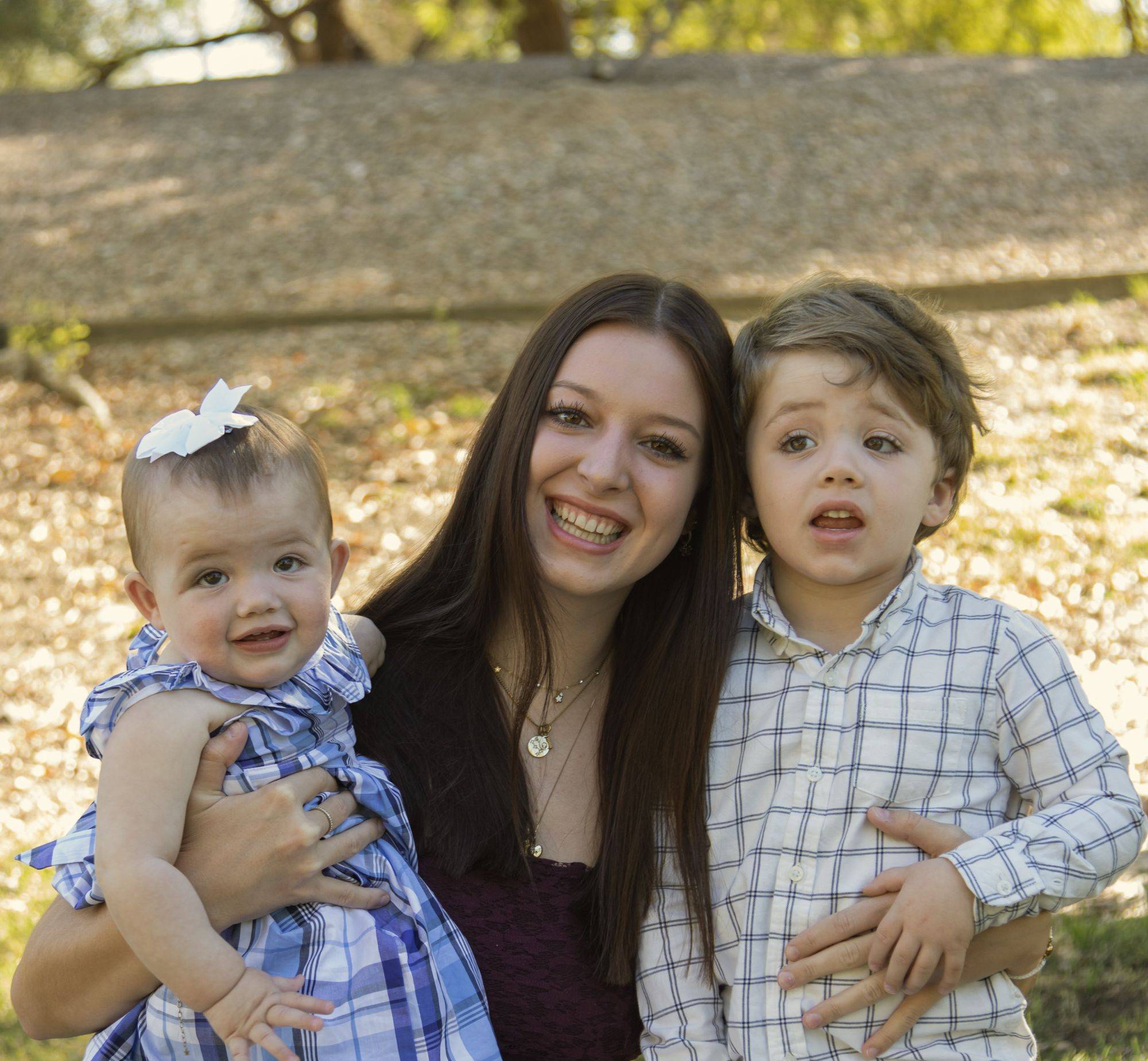 A woman is smiling, holding a baby girl and standing next to a young boy, all outdoors in a park. The children are wearing plaid outfits.