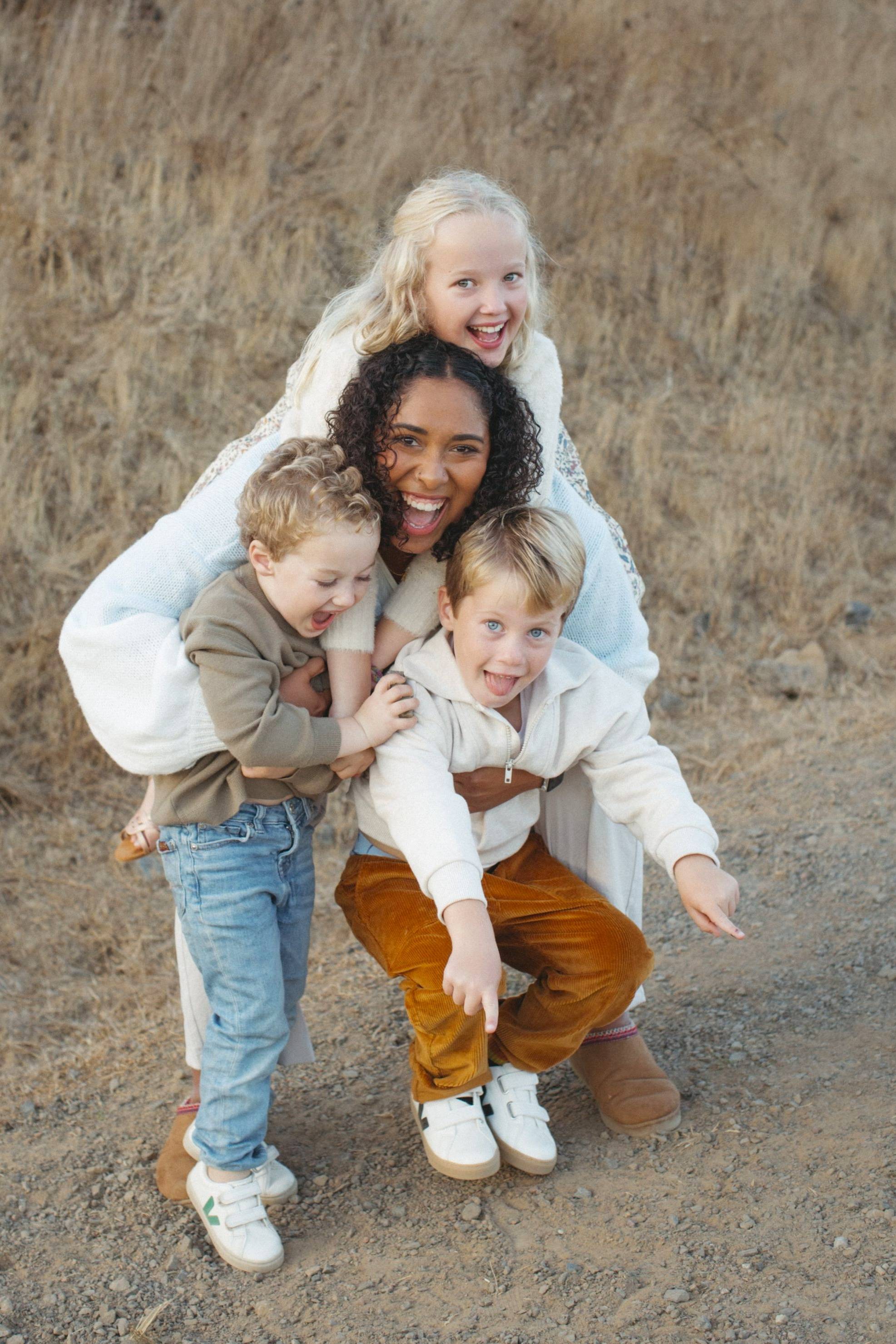 A smiling woman crouches down with three joyful children draped over her as they play outdoors on a dirt path.