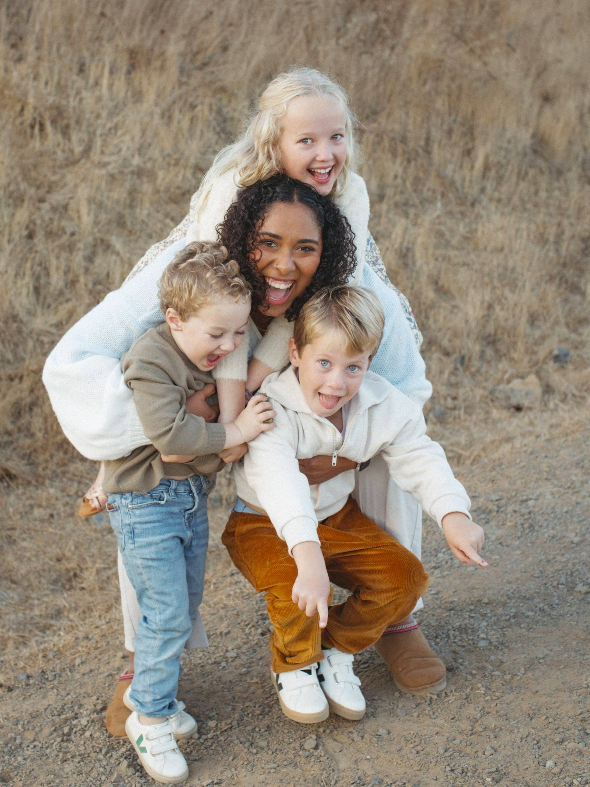 A smiling woman crouches down with three joyful children draped over her as they play outdoors on a dirt path.
