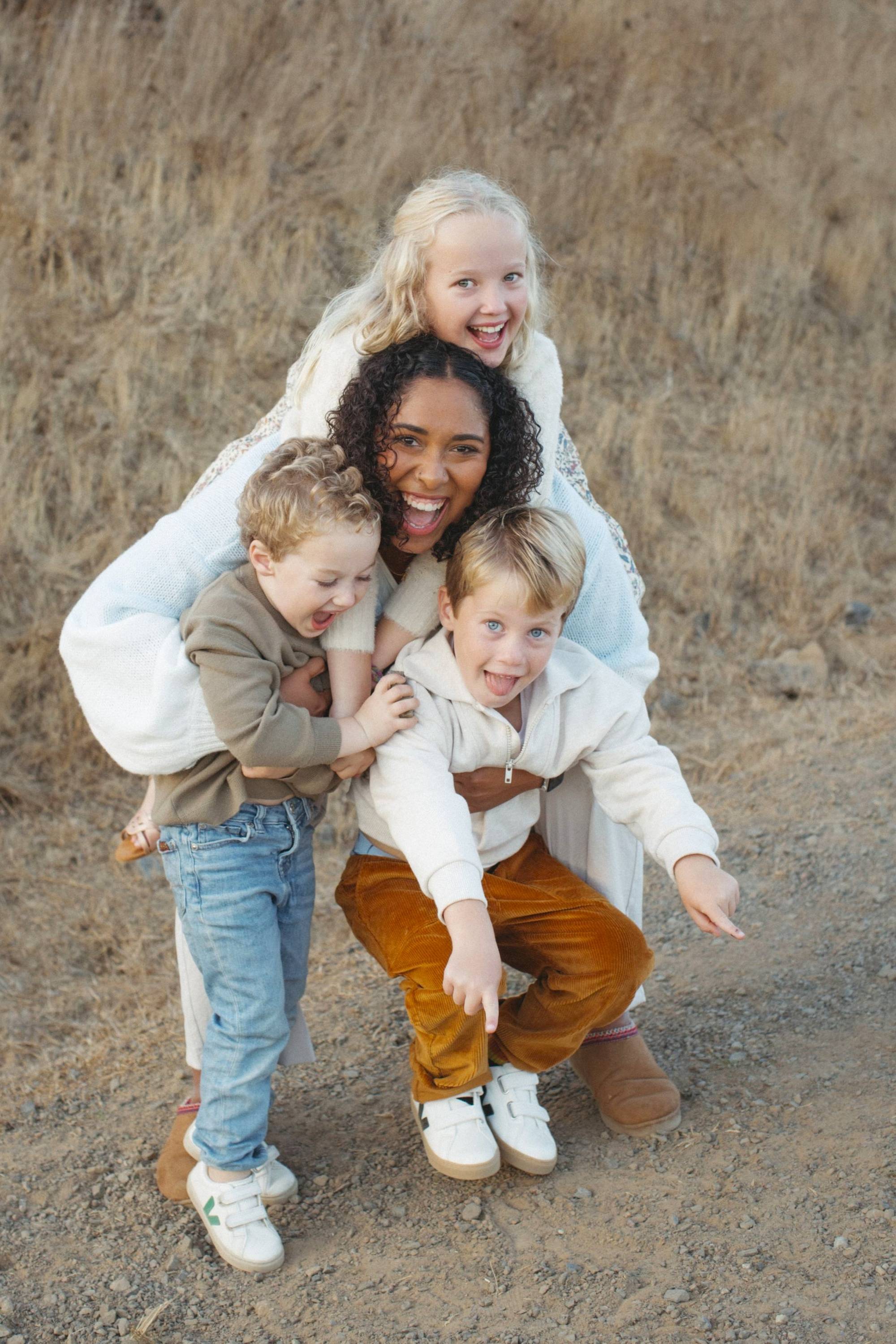 A smiling woman crouches down with three joyful children draped over her as they play outdoors on a dirt path.