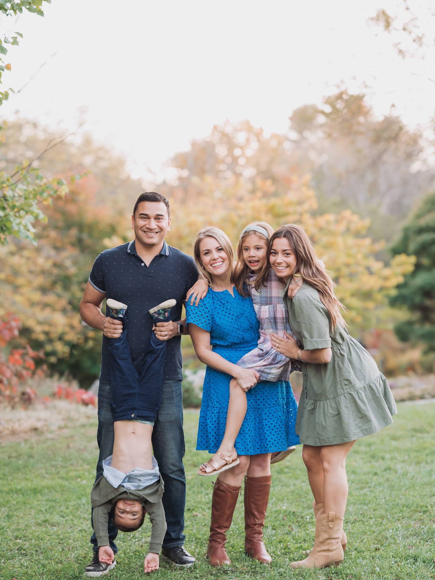 A family poses outdoors; a man playfully holds a child upside down, while two women and a girl smile. Autumn foliage is in the background.