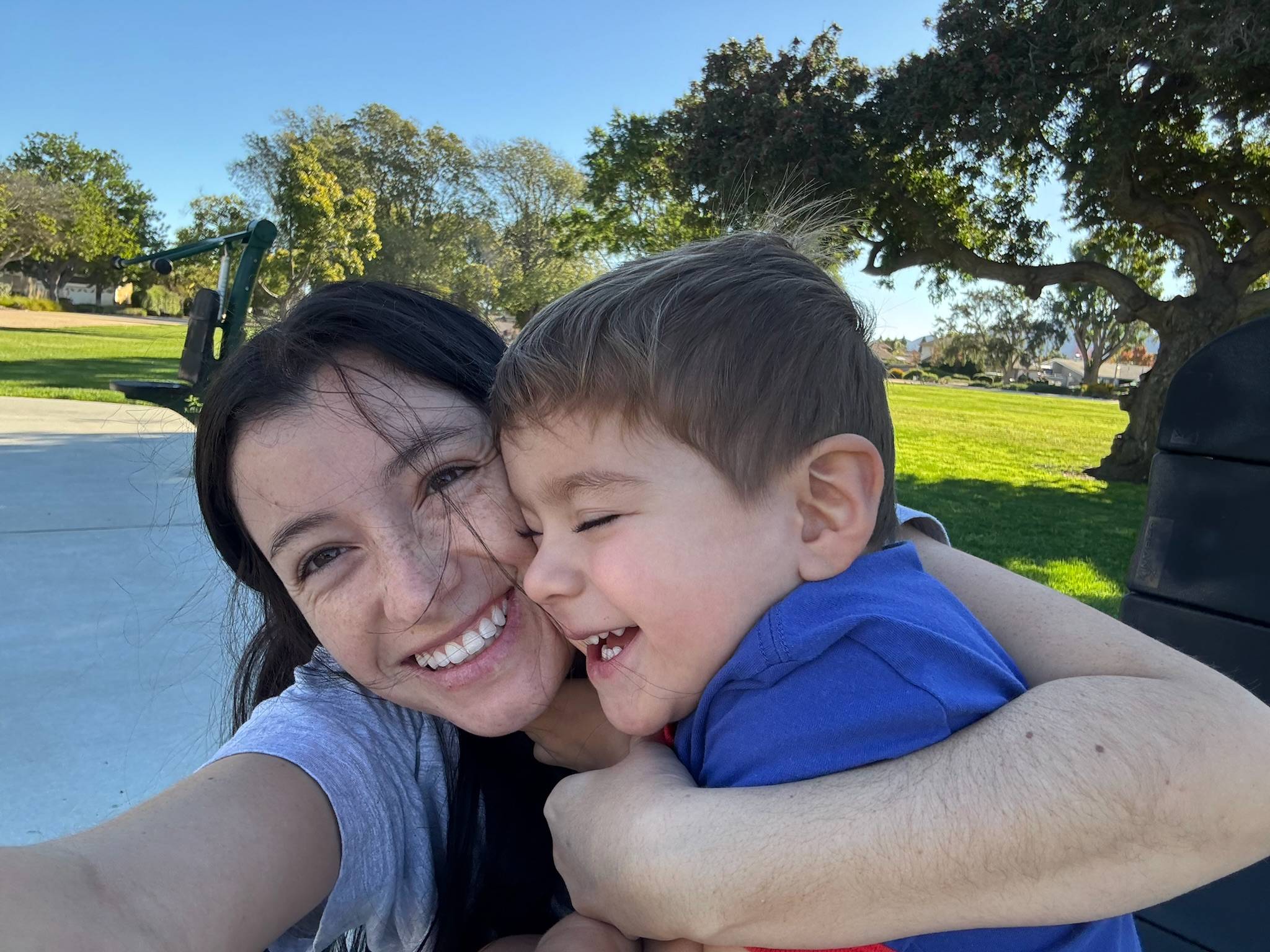 A woman and a child share a joyful hug outdoors in a sunny park, with trees and grass in the background. Both are smiling brightly.
