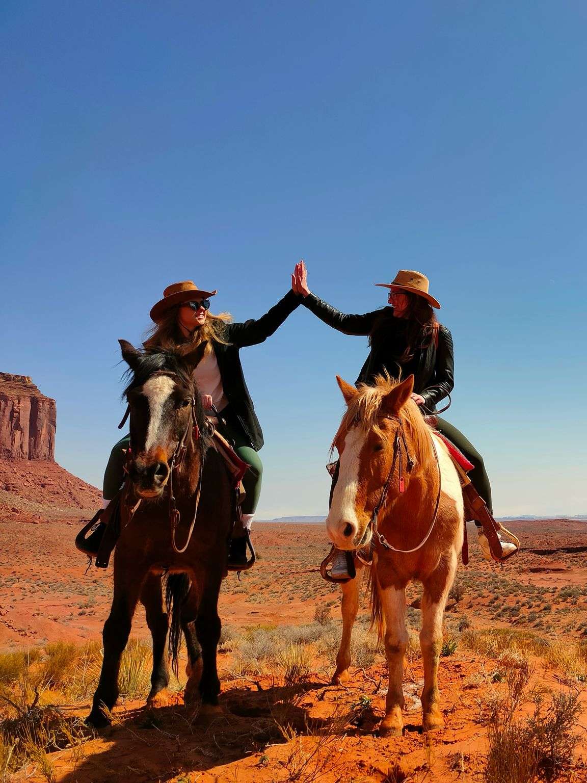 Two people on horseback high-five in a desert landscape with red rock formations and a clear blue sky.