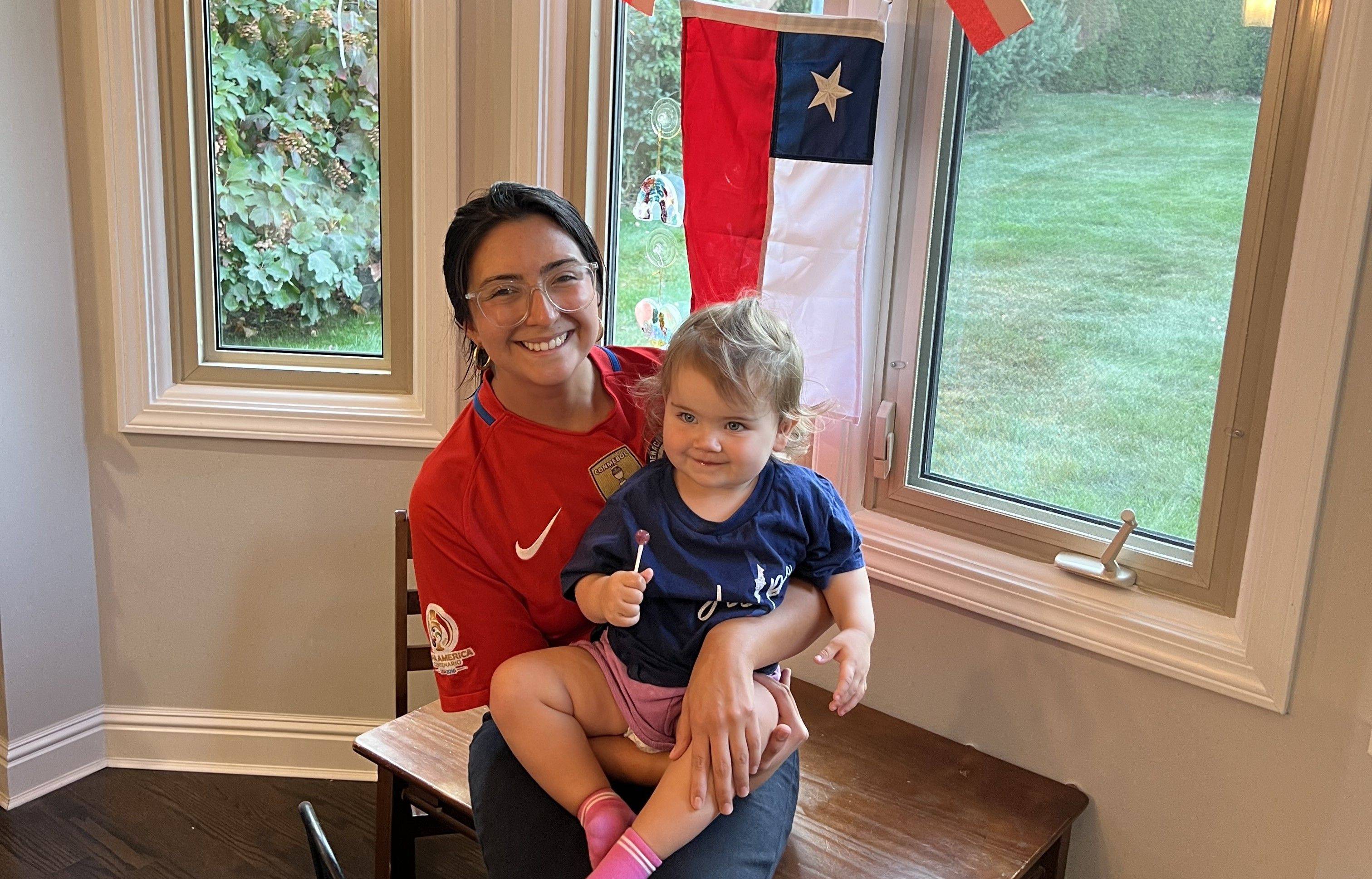 Person holding a toddler on their lap, sitting by a window with a Chilean flag hanging. Greenery is visible outside. Both are smiling.