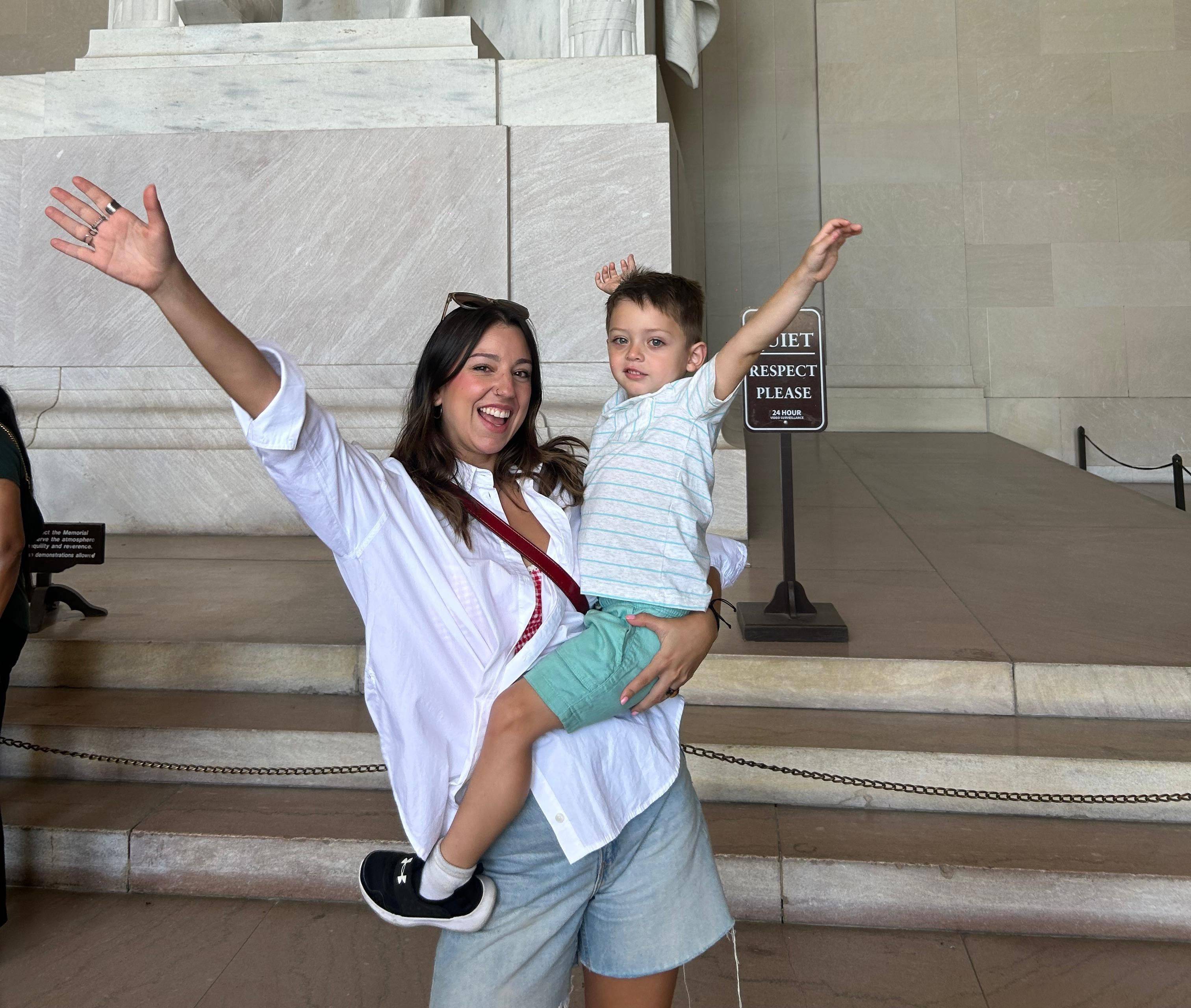 A woman holds a child, both smiling with raised arms, in front of a large marble structure. A sign in the background reads "Respect Please."